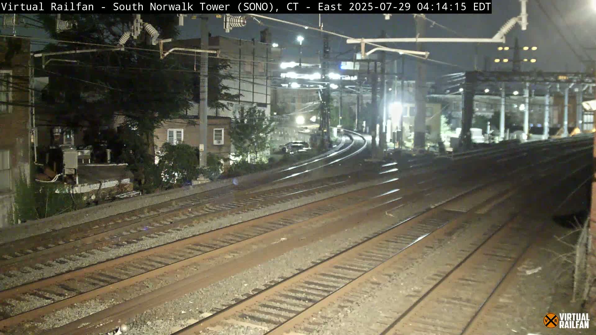 Multiple train tracks extend into the distance at night, curving slightly to the right past buildings and trees under artificial lighting.