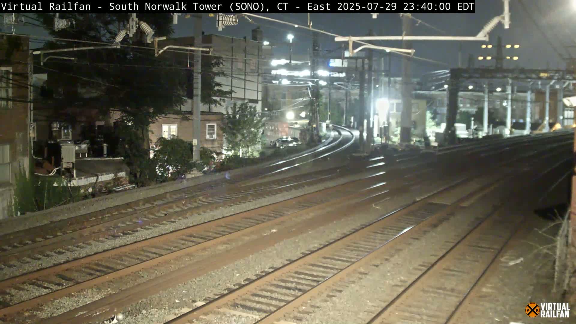 Multiple train tracks extend into the distance at night, curving slightly to the right past buildings and trees under artificial lighting.