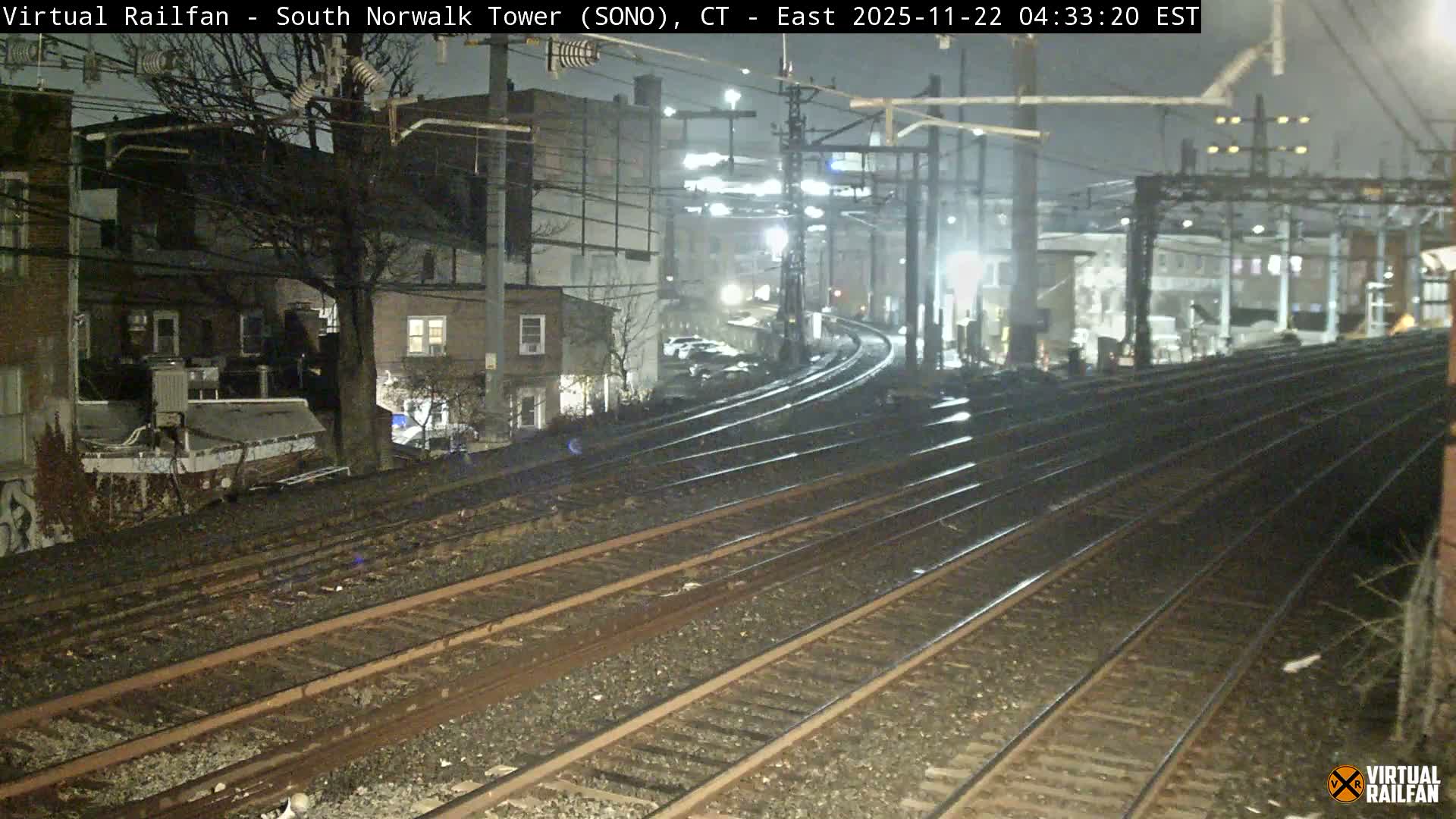 Multiple railroad tracks curve into the distance past various buildings with lit windows and overhead electrical structures under a clear night sky.