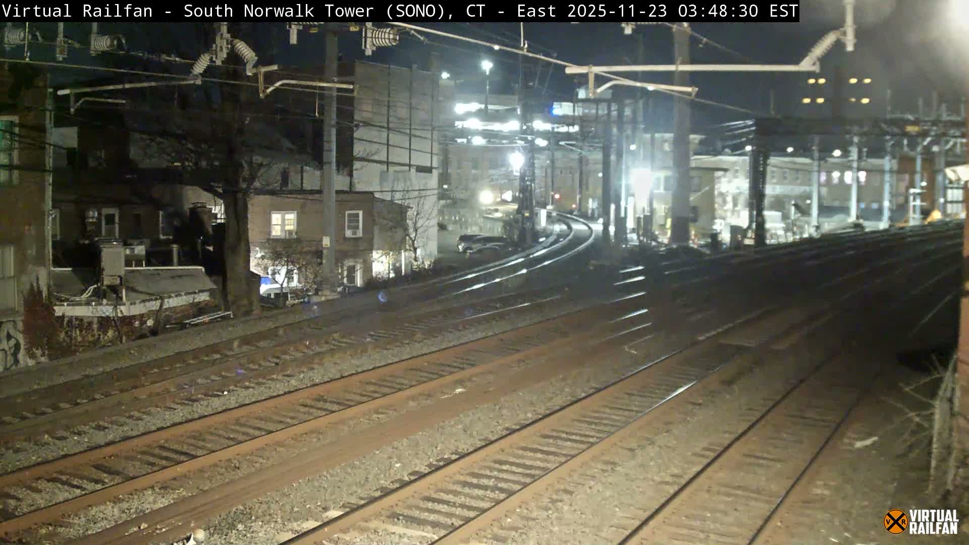 A clear night view captures multiple railroad tracks curving through an urban landscape, illuminated by overhead streetlights and the lights of nearby buildings with bare trees scattered between them.