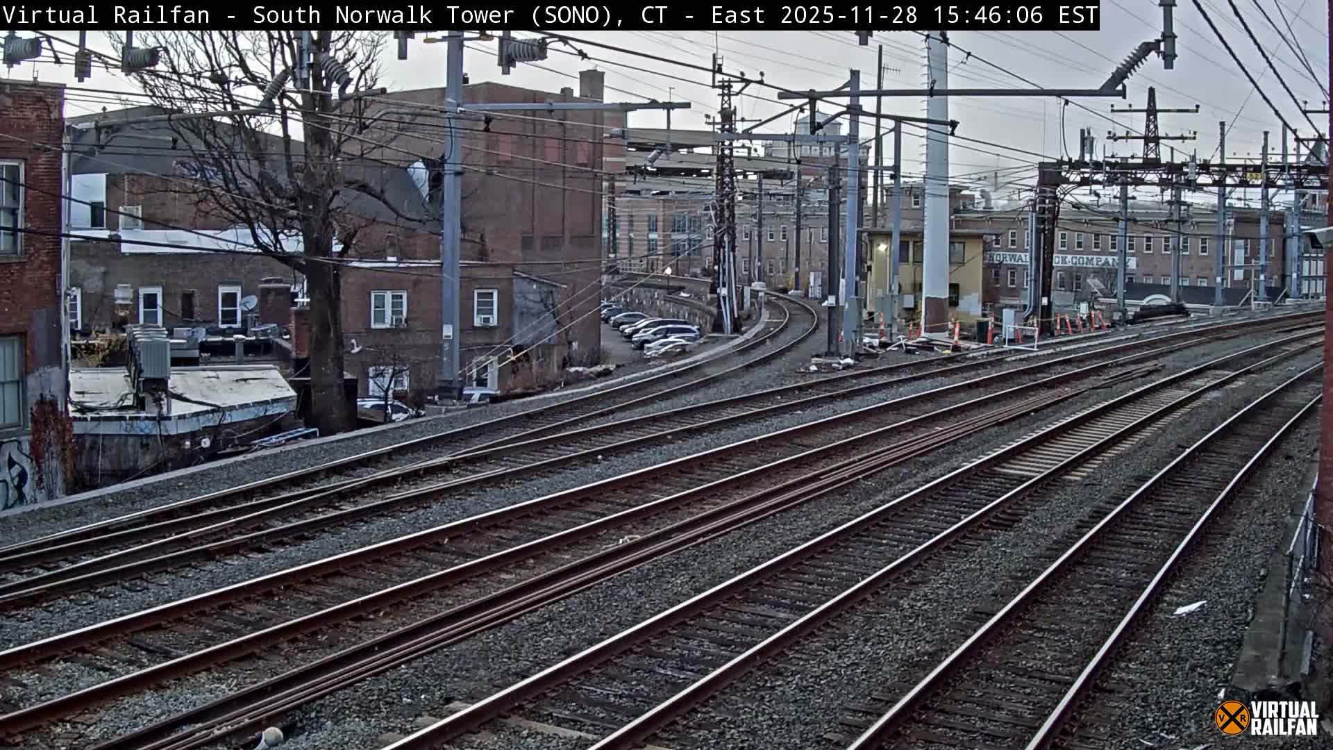 The image captures a wide view of numerous parallel railroad tracks curving into the distance amidst an urban landscape of buildings and bare trees, under an overcast sky.