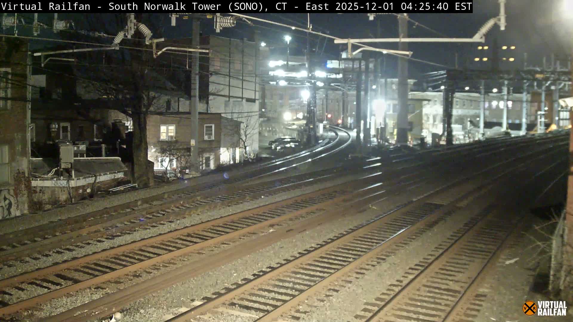 Multiple illuminated train tracks curve through an urban landscape at night, flanked by various buildings and bare trees under clear, dry conditions.