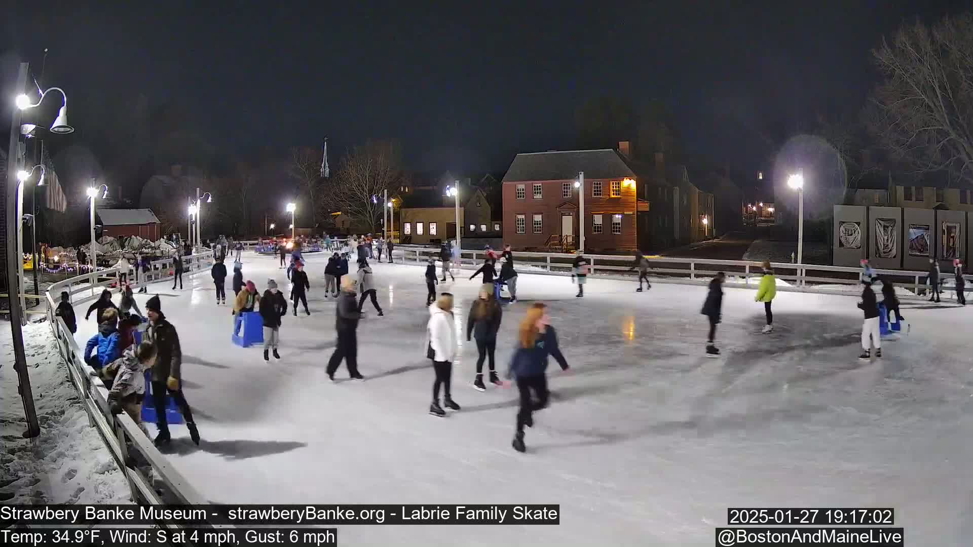 Many people ice skate at night on an outdoor rink, illuminated by lights, near buildings.