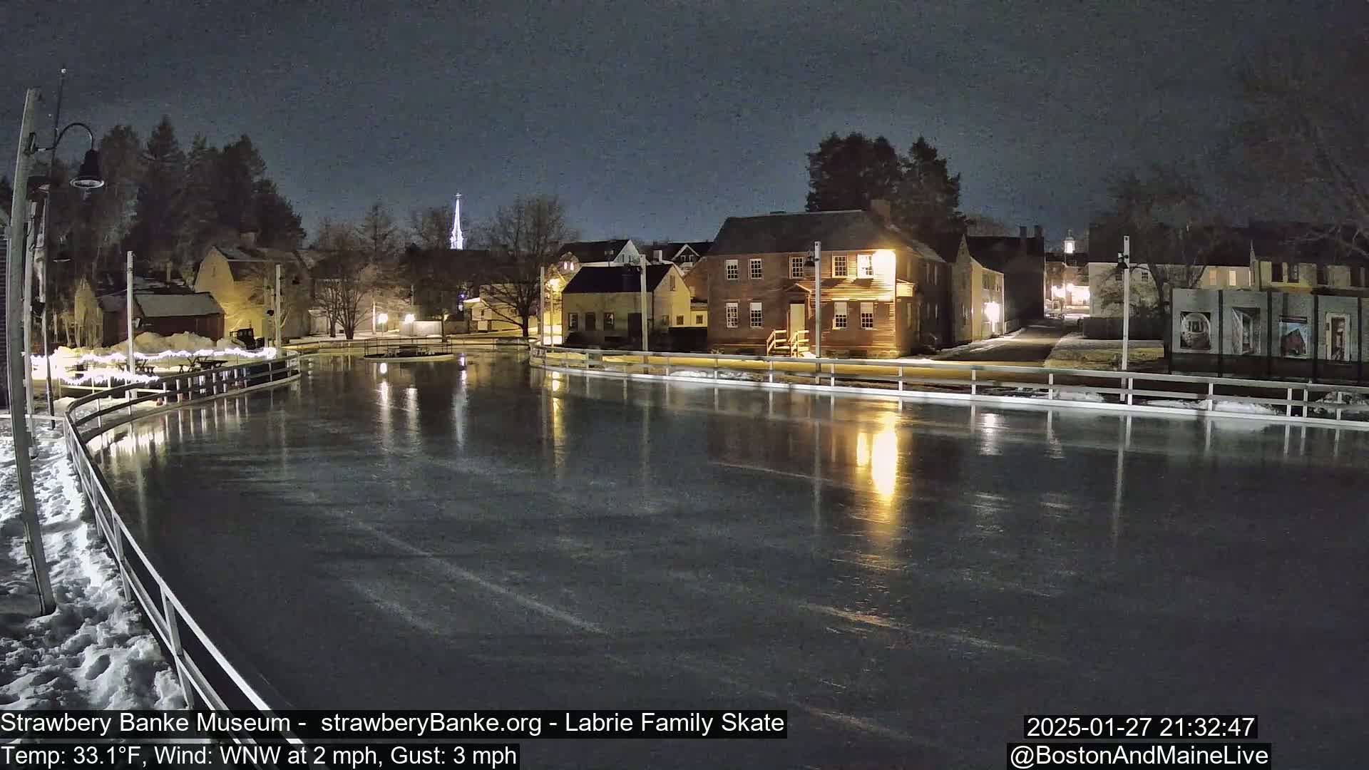 A nighttime view of an outdoor ice skating rink illuminated by nearby buildings and streetlights, with a light wind.