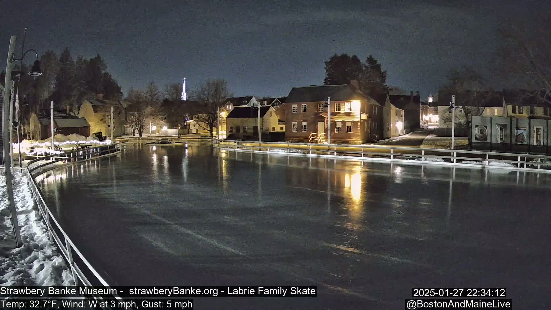 A nighttime view of an outdoor ice skating rink, illuminated by building lights, surrounded by snow-covered ground and historic-looking buildings under a dark sky.