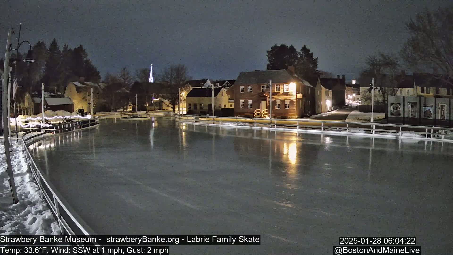 A nighttime view of an outdoor ice rink, illuminated by nearby buildings, under a dark sky with a temperature of 33.6°F and a southwest wind.