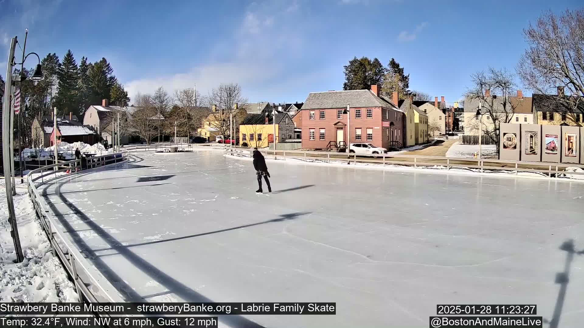 A person ice skates on an outdoor rink in front of historic buildings on a sunny day with a northwest wind.