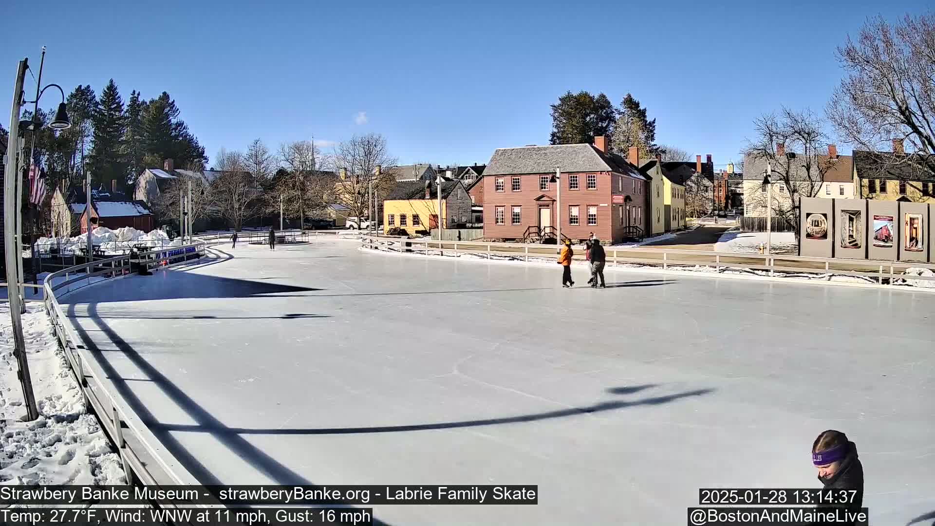 A sunny, outdoor ice rink with several people ice skating in front of historic buildings, under a clear blue sky.