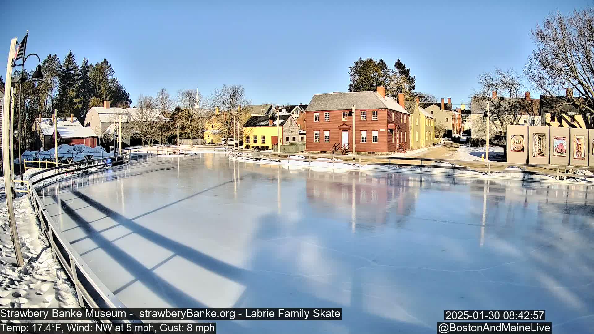 A frozen skating rink reflects nearby colonial-style buildings under a clear, sunny sky.