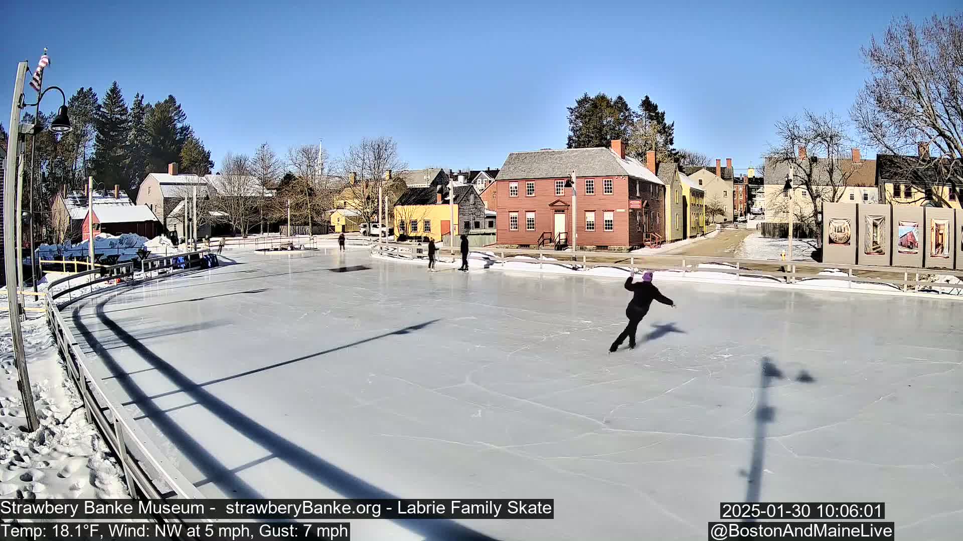 Several people ice skate on an outdoor rink on a sunny day with a northwest wind.