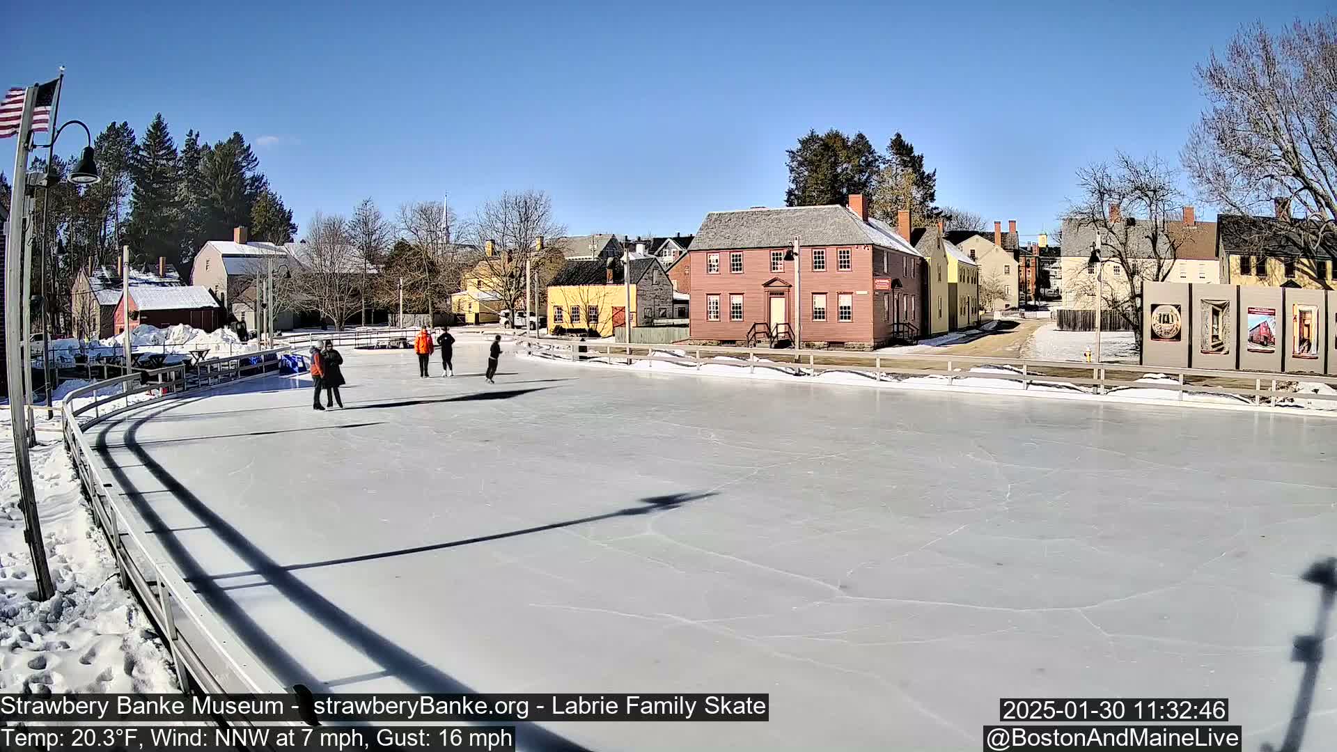 A sunny, outdoor ice skating rink with several people skating, situated near historic-looking buildings and trees.