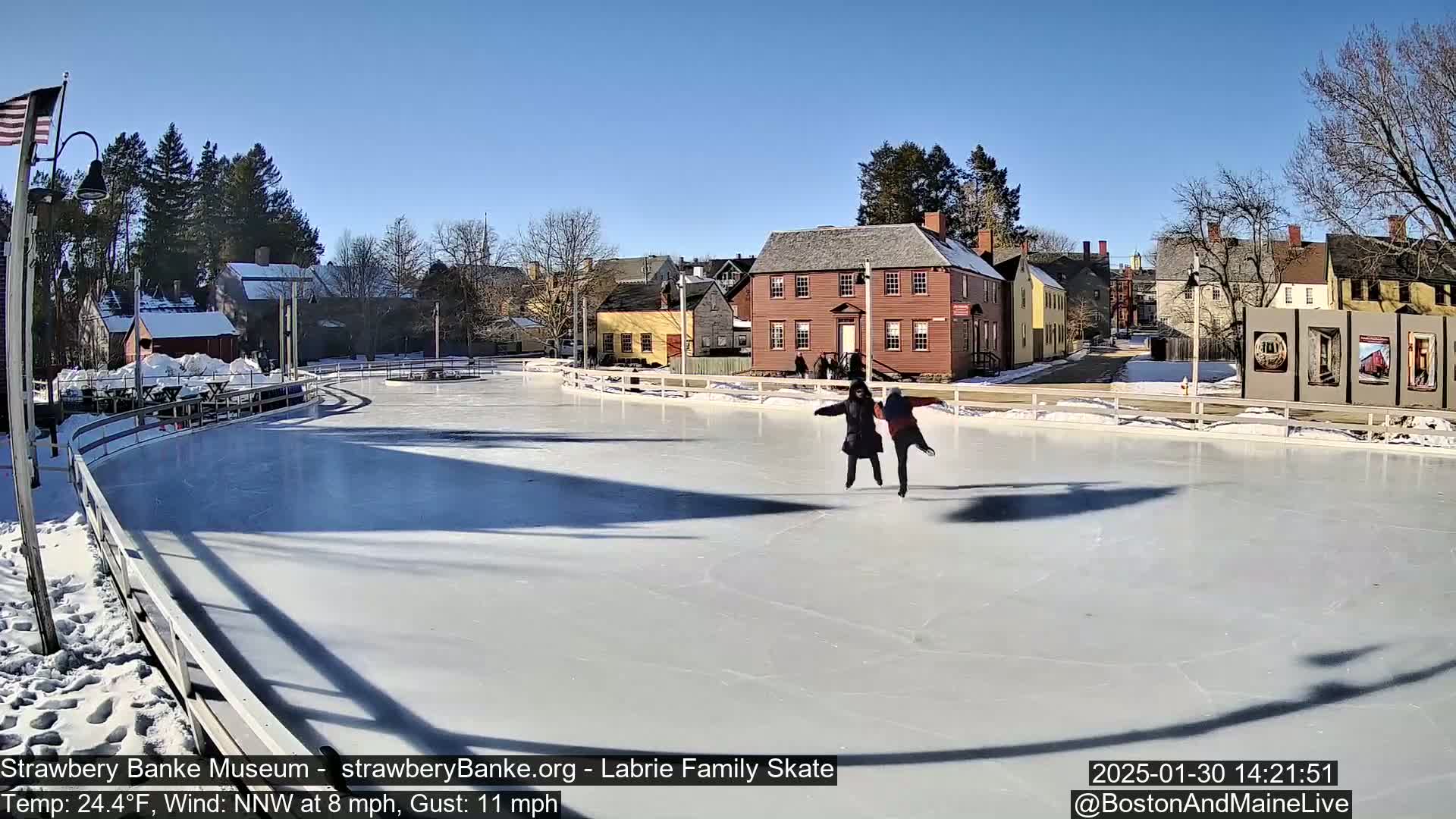 Two people ice skate on an outdoor rink on a sunny day with a light wind.