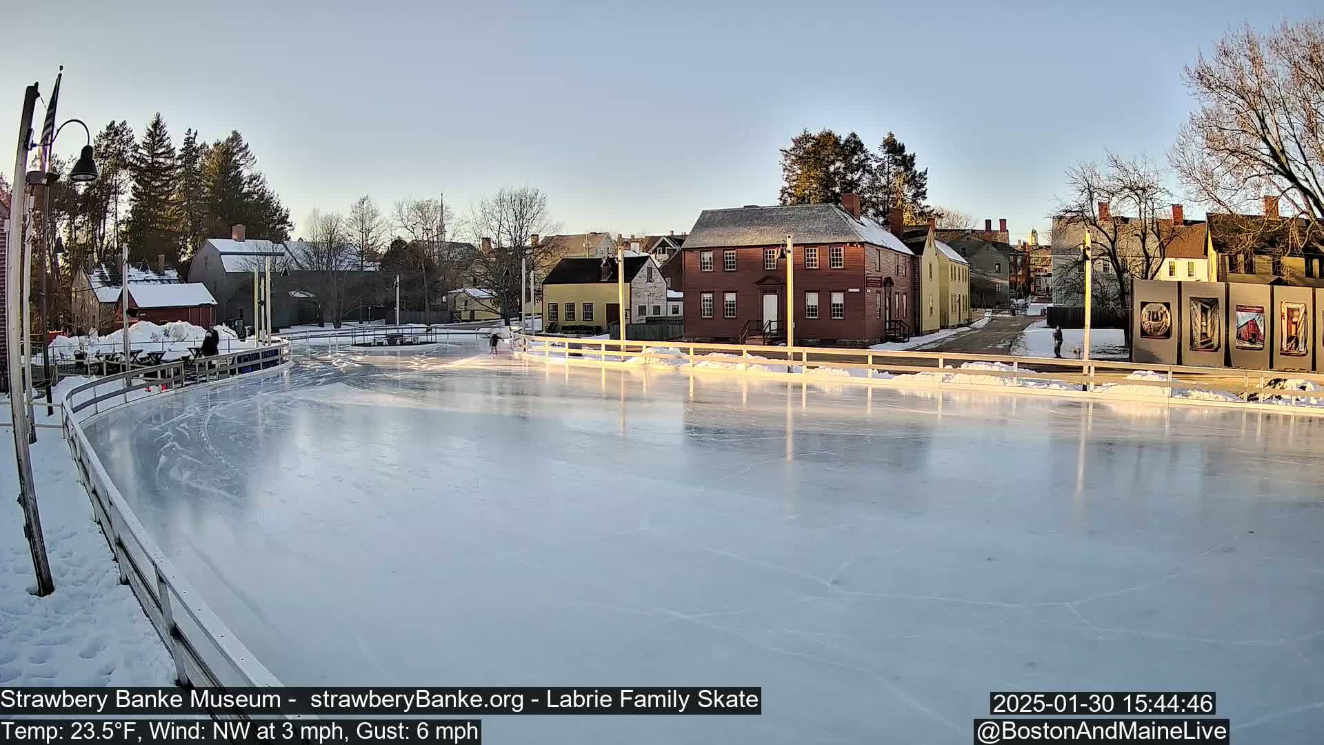 A frozen outdoor skating rink is surrounded by historic-looking buildings under a clear, sunny sky.