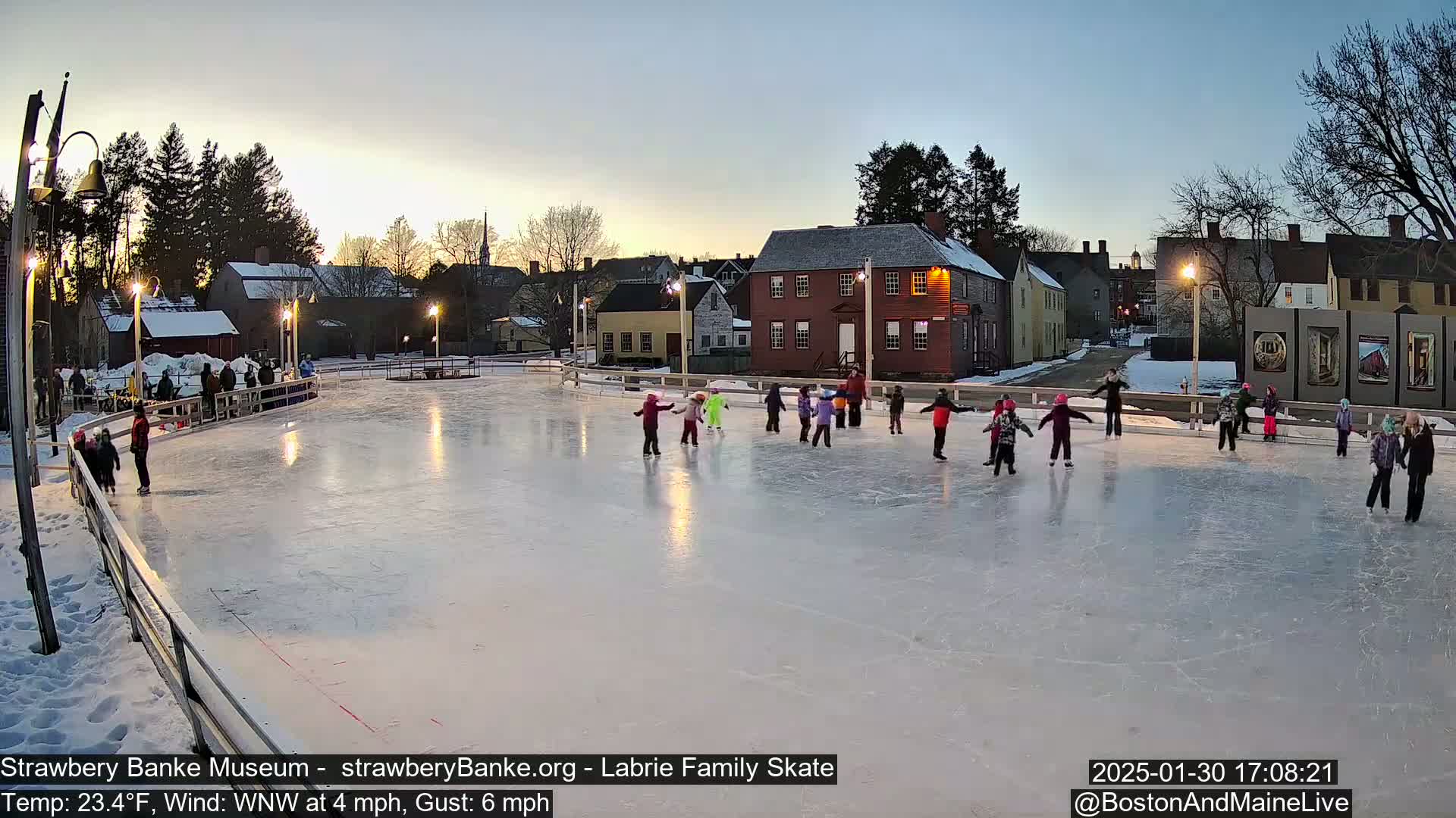 Many children ice skate on an outdoor rink at dusk, under a clear sky with a light wind.