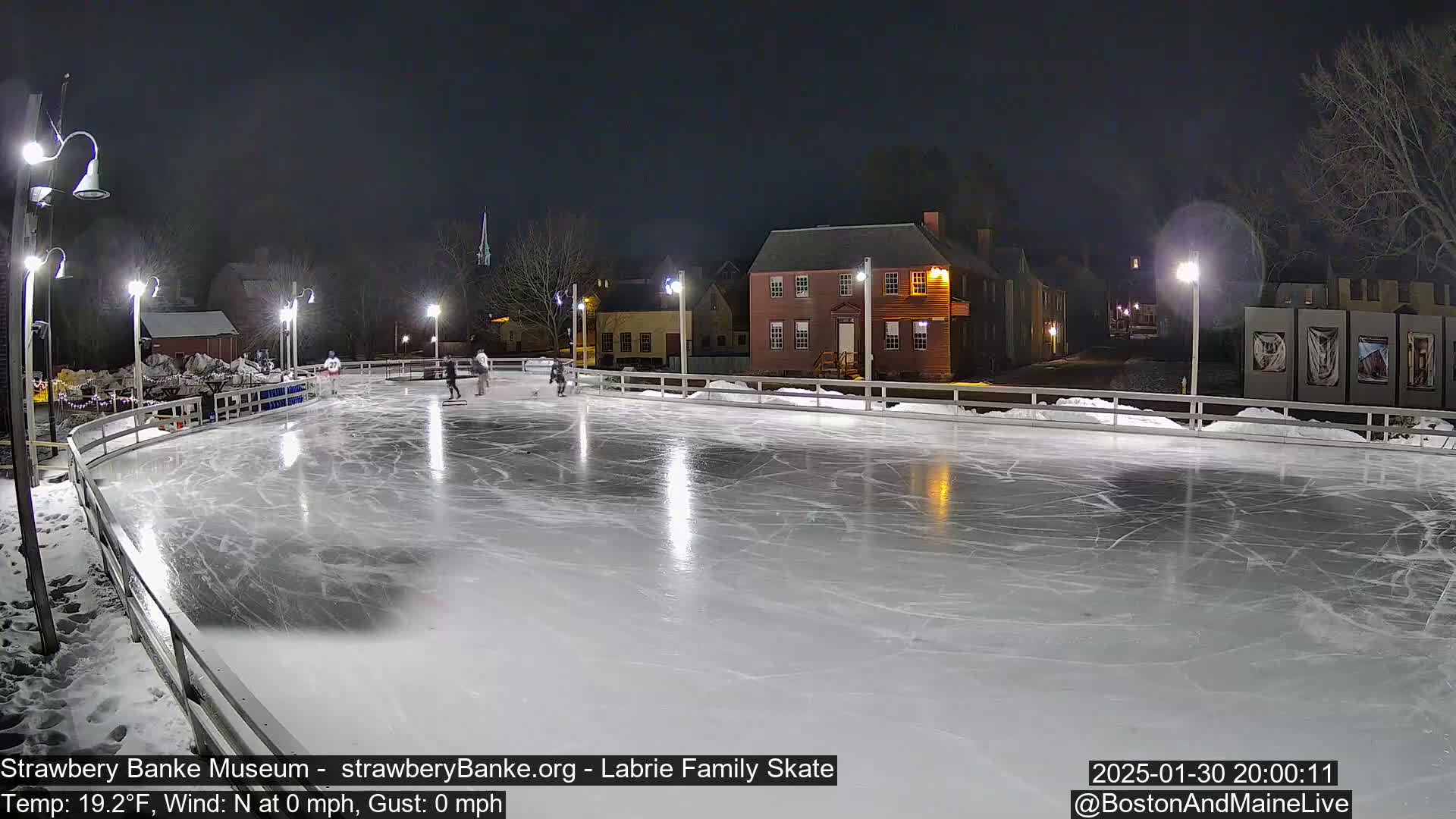 A brightly lit outdoor ice rink at night with several people ice skating, surrounded by historic buildings and trees under a clear, dark sky.