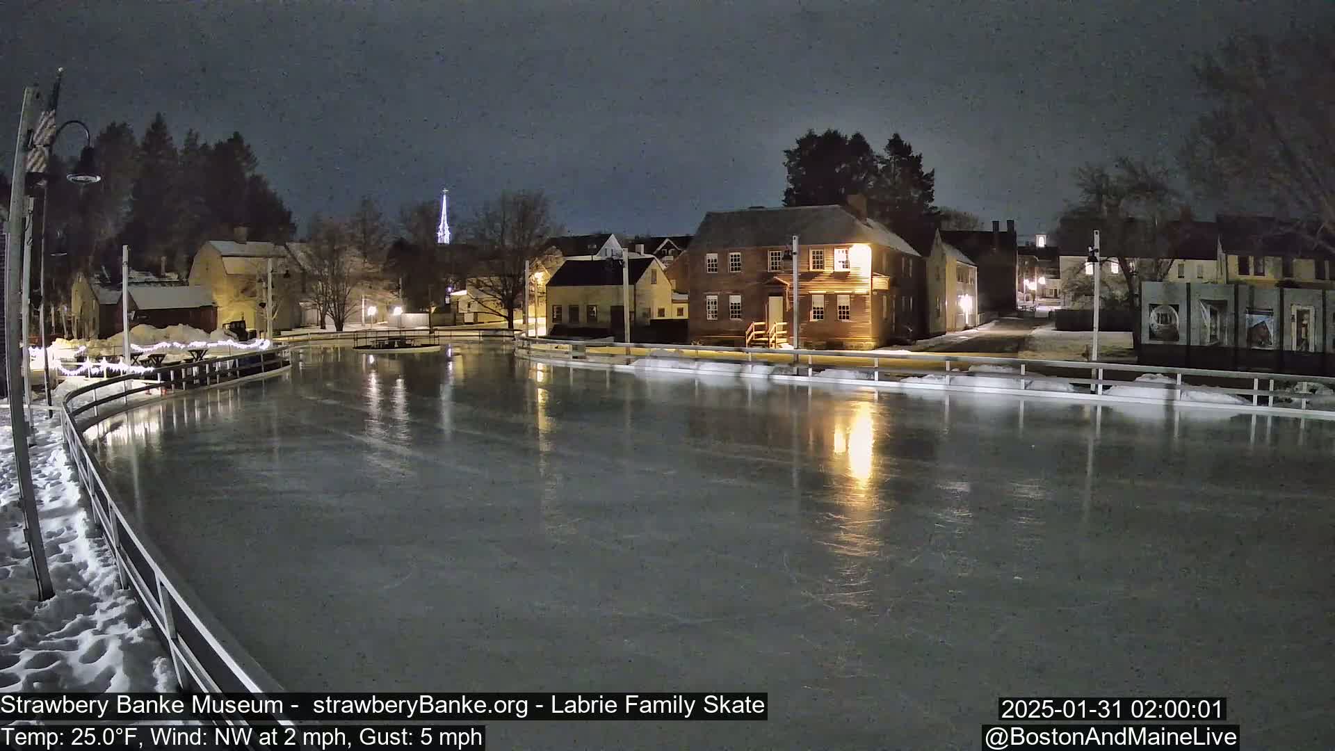 A nighttime view of an ice skating rink illuminated by lights, surrounded by snow-covered buildings under a dark sky.