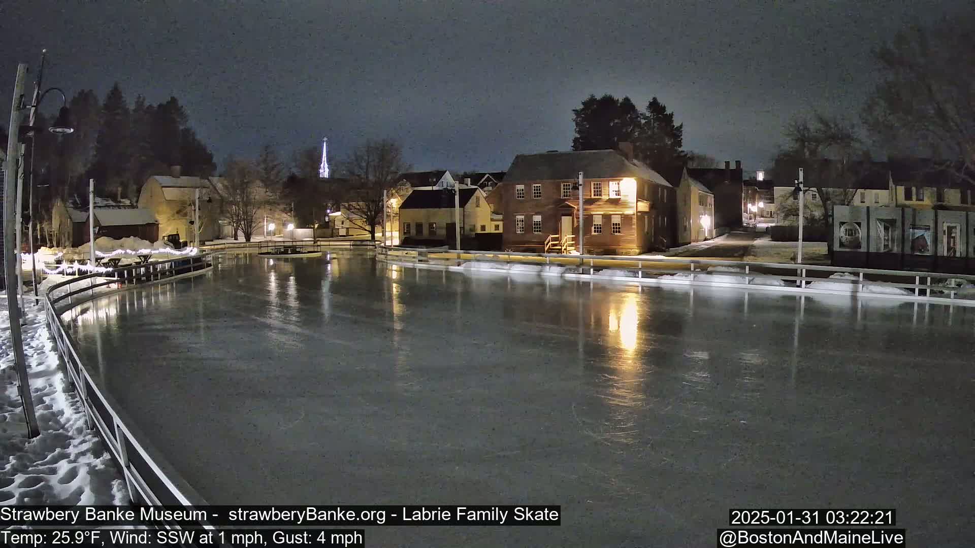 A nighttime view of a snow-covered, ice-skating rink illuminated by lights, reflecting the nearby buildings and a church steeple under a dark sky.