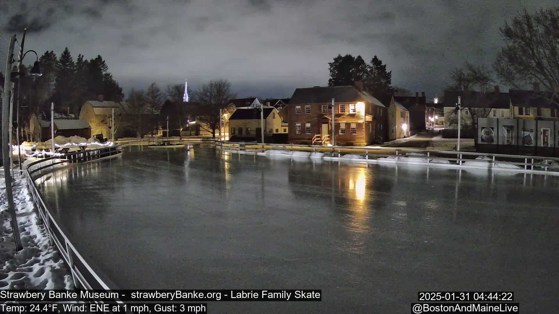 A nighttime view of an outdoor ice skating rink, illuminated by lights, next to historic-looking buildings under a dark, cloudy sky.