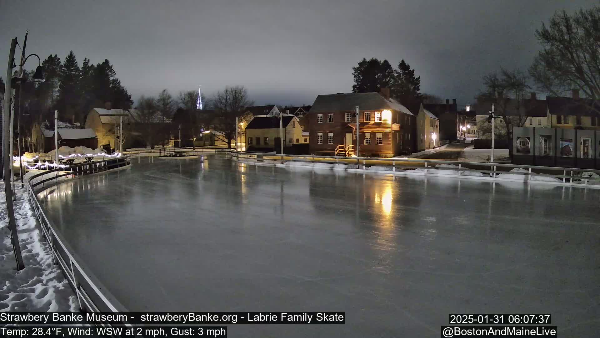 A nighttime view of an outdoor ice skating rink, illuminated by nearby lights, with a small town and a church steeple visible in the background under a dark, overcast sky.