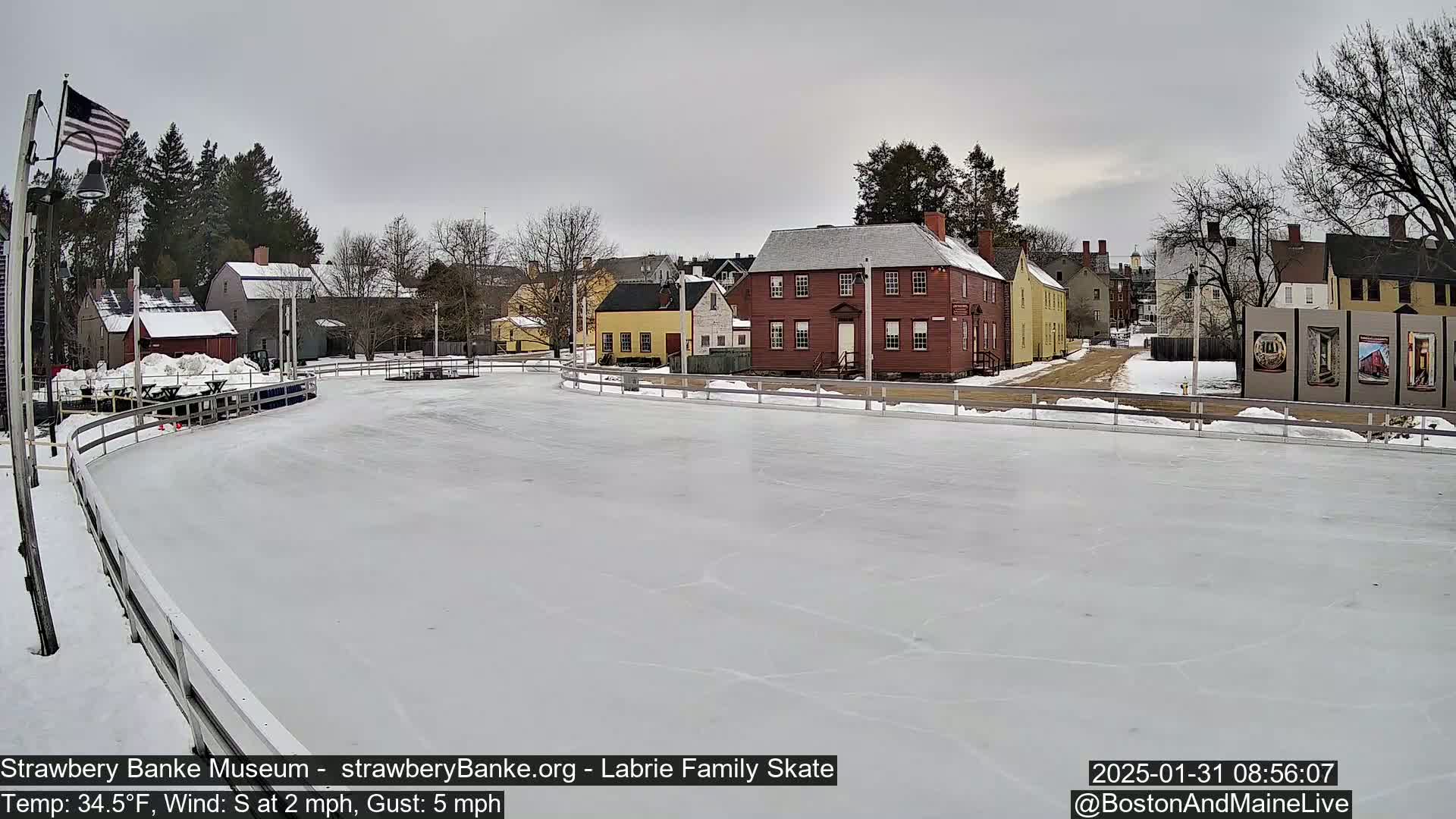 A snow-covered outdoor ice rink sits in front of several historic-looking buildings under an overcast sky.