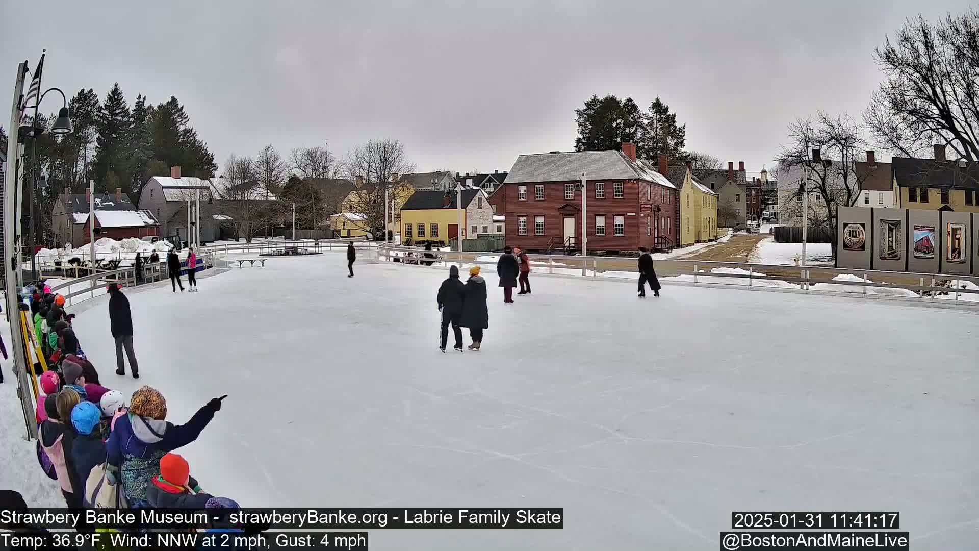 An outdoor ice skating rink, surrounded by historical-looking buildings and snow-covered ground, shows several people ice skating and others watching under an overcast sky.