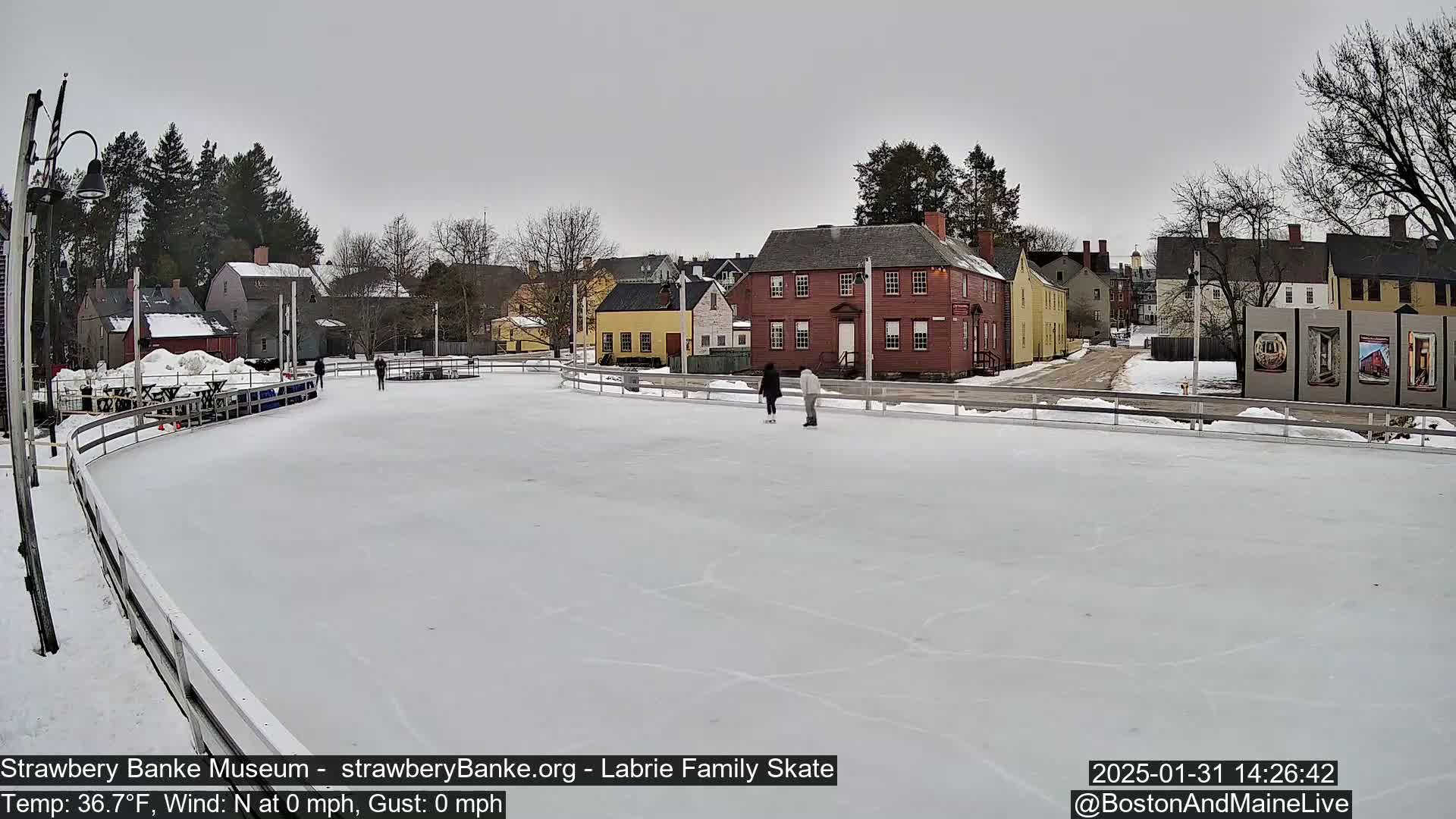 Two people ice skate on a rink in front of several historic-looking buildings on an overcast day.
