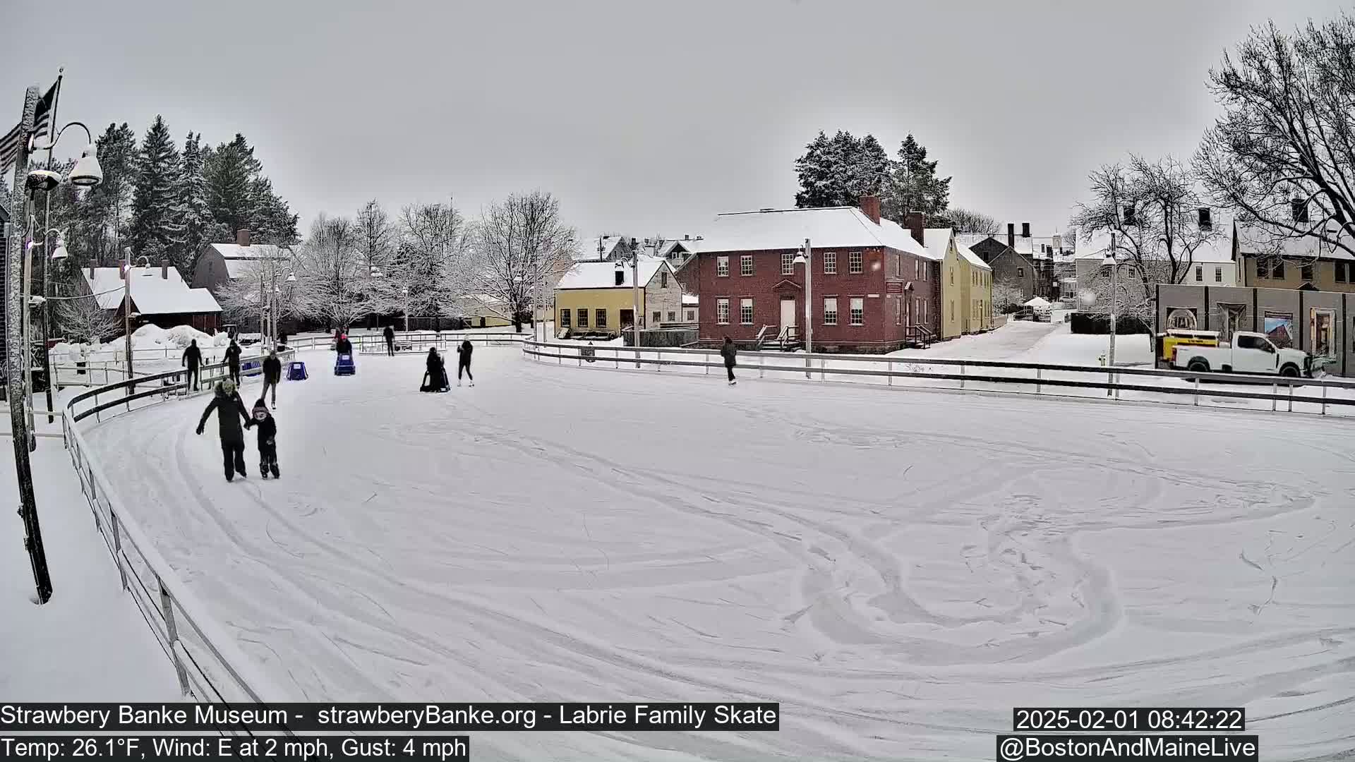 People ice skate on an outdoor rink surrounded by snow-covered buildings under an overcast sky.