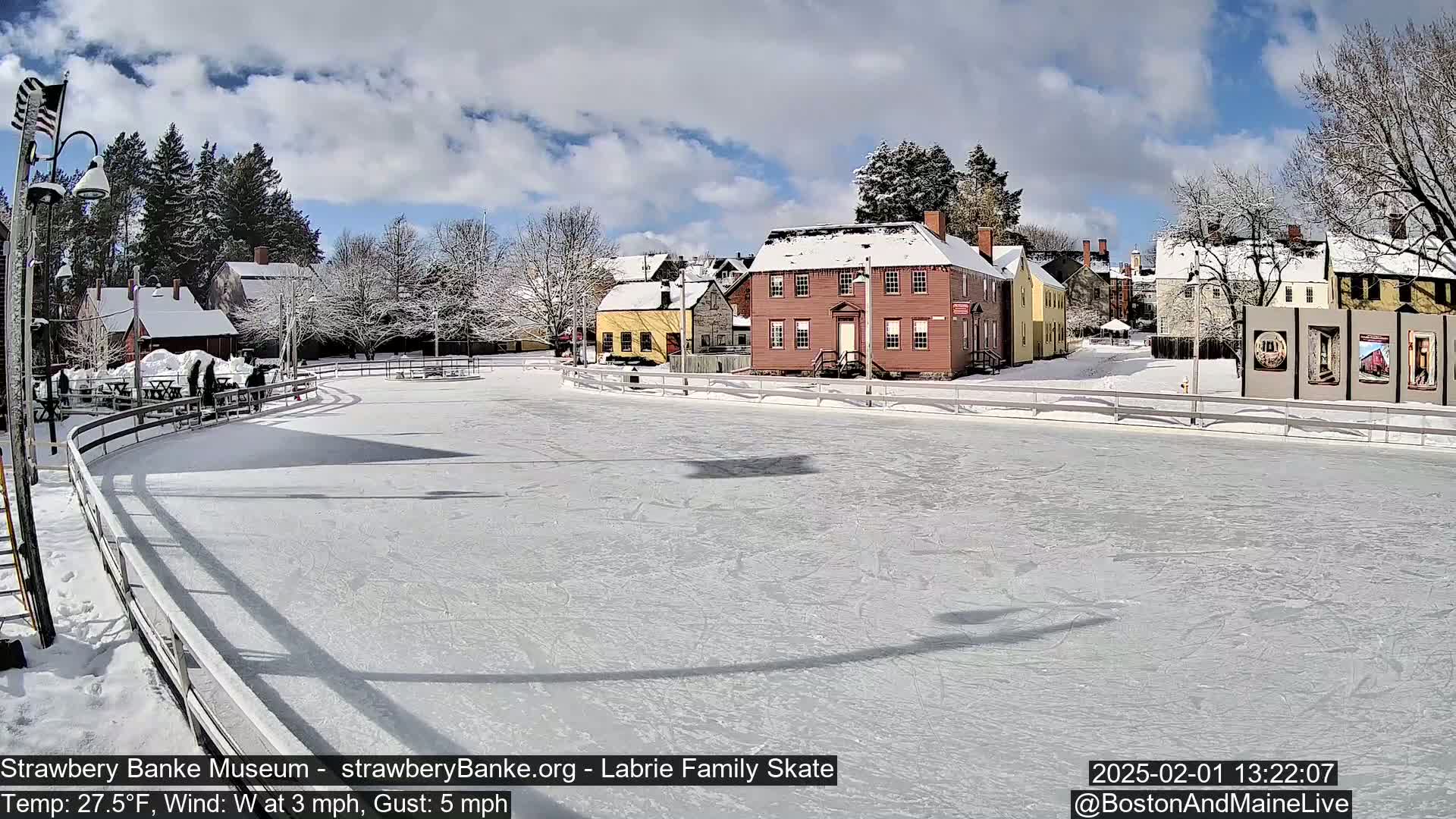 A snow-covered outdoor ice skating rink is situated in front of several historic-looking buildings under a partly cloudy sky.