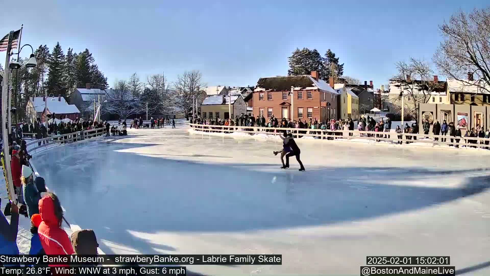 A pair of figure skaters glides across an outdoor ice rink surrounded by spectators on a sunny day with a light WNW wind.
