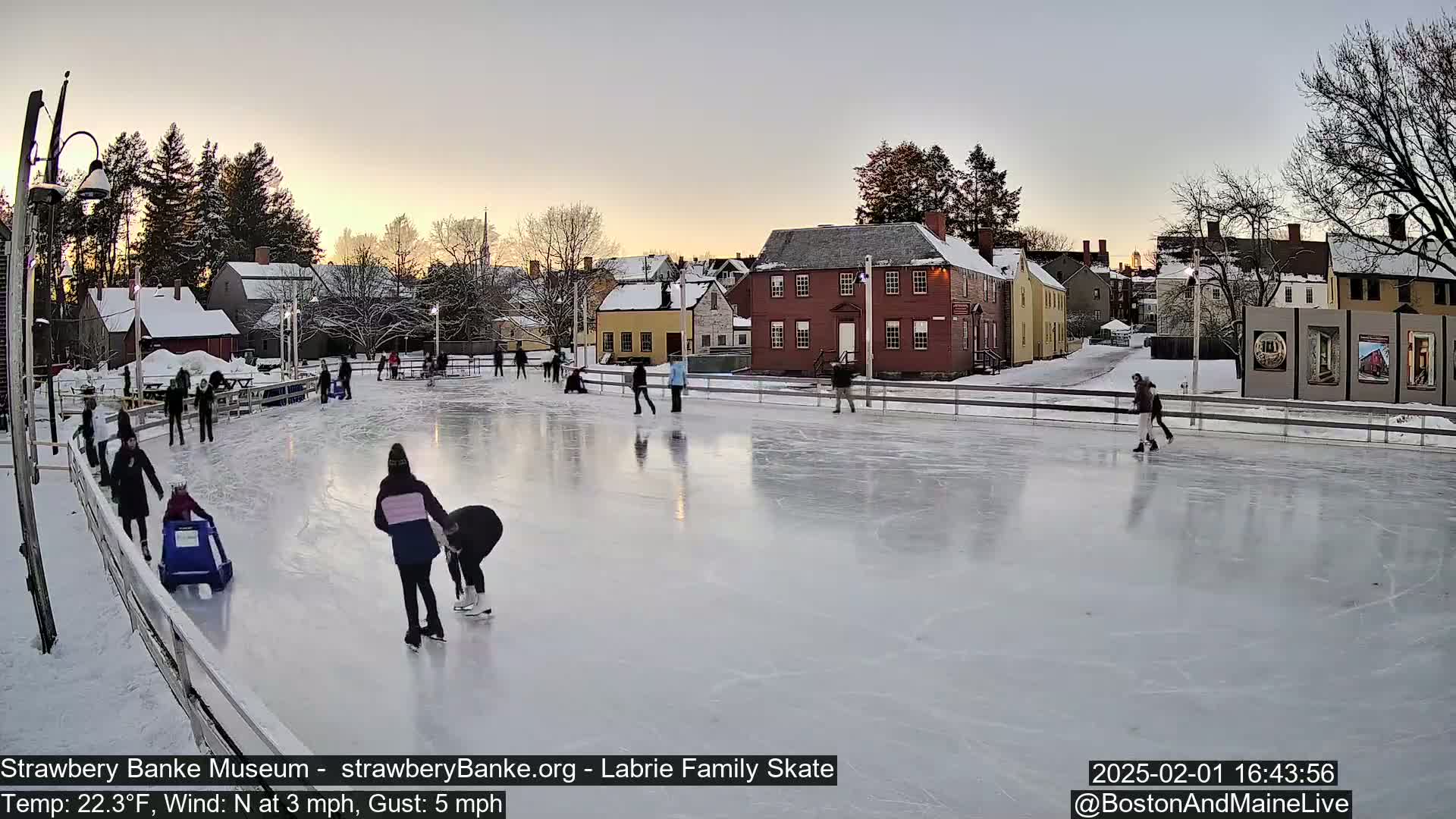 People ice skate on an outdoor rink at sunset, surrounded by snow-covered buildings and trees in a cold, windy setting.