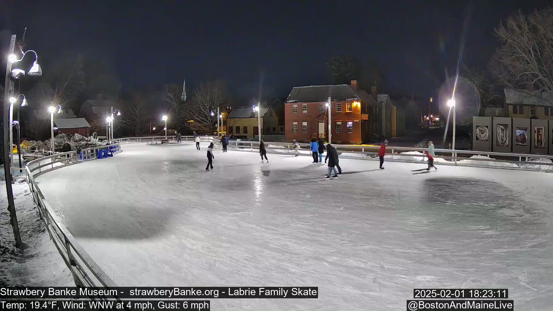 Several people ice skate on an outdoor rink at night under lights, with a light wind.