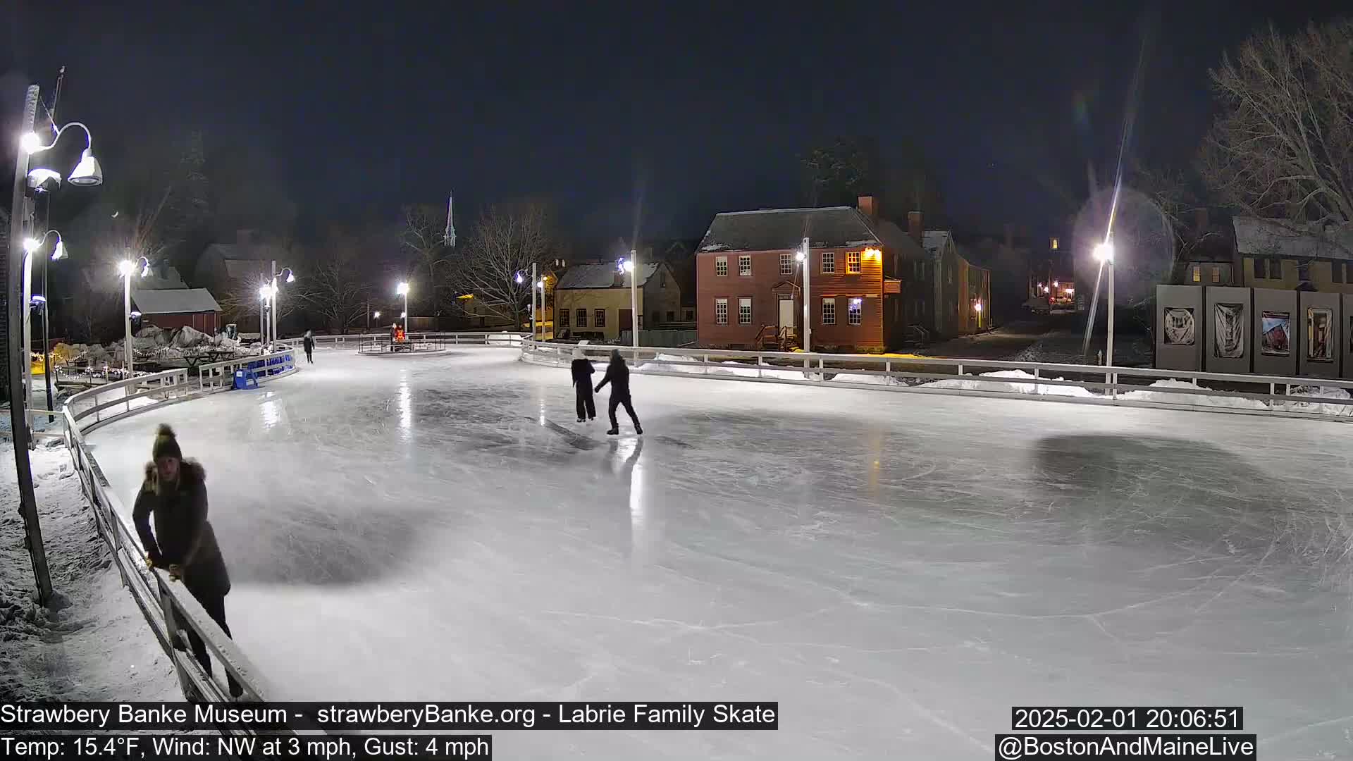 At night, three people ice skate on an outdoor rink illuminated by lights, surrounded by snow-covered buildings under a dark sky.