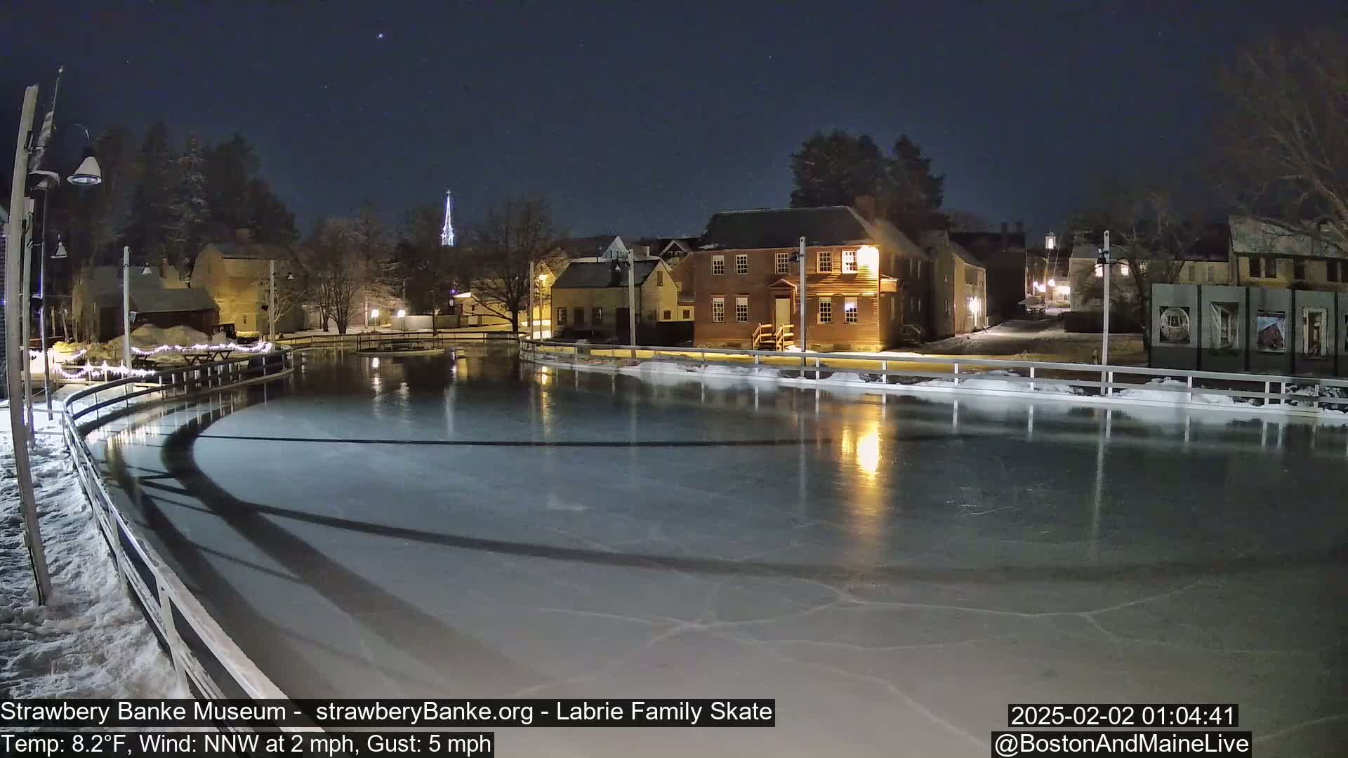 A nighttime view of an ice skating rink, illuminated, surrounded by historic-looking buildings under a clear, dark sky.
