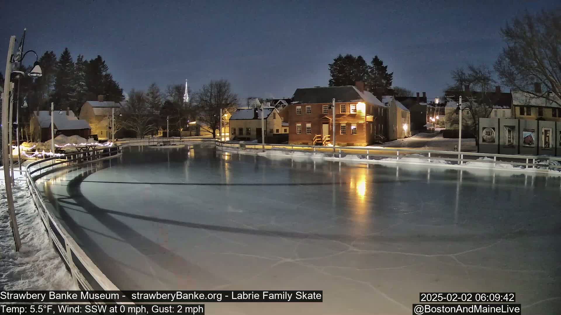 An outdoor ice skating rink, illuminated at night under a dark sky, is situated in front of historic-looking buildings.