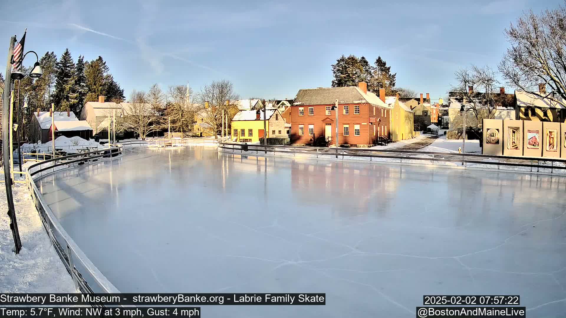 A frozen outdoor skating rink, surrounded by snow-covered colonial-style buildings under a clear, sunny sky.
