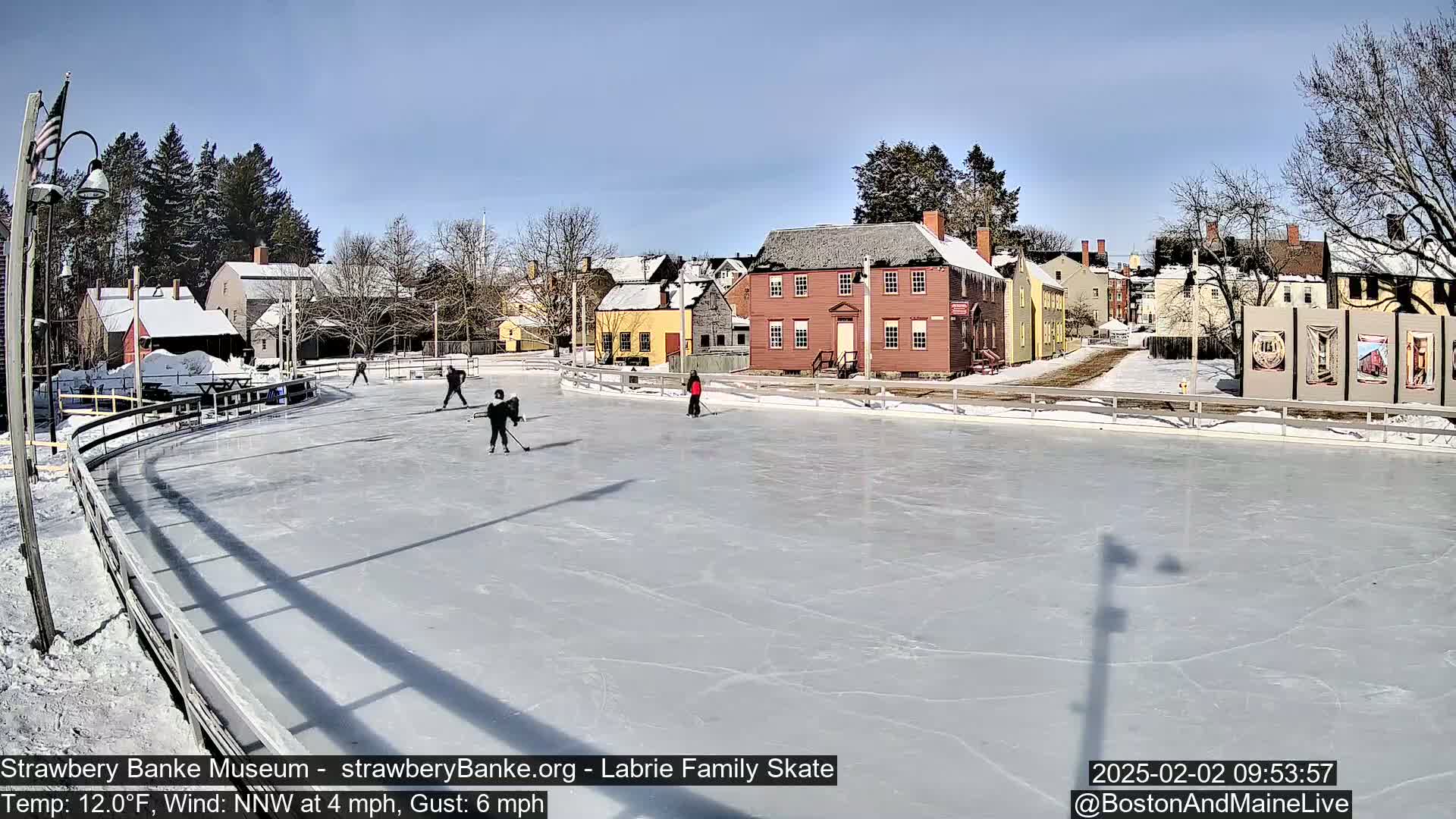 Several people ice skate on an outdoor rink in front of historic-looking buildings on a sunny, cold day with a light north-northwest wind.