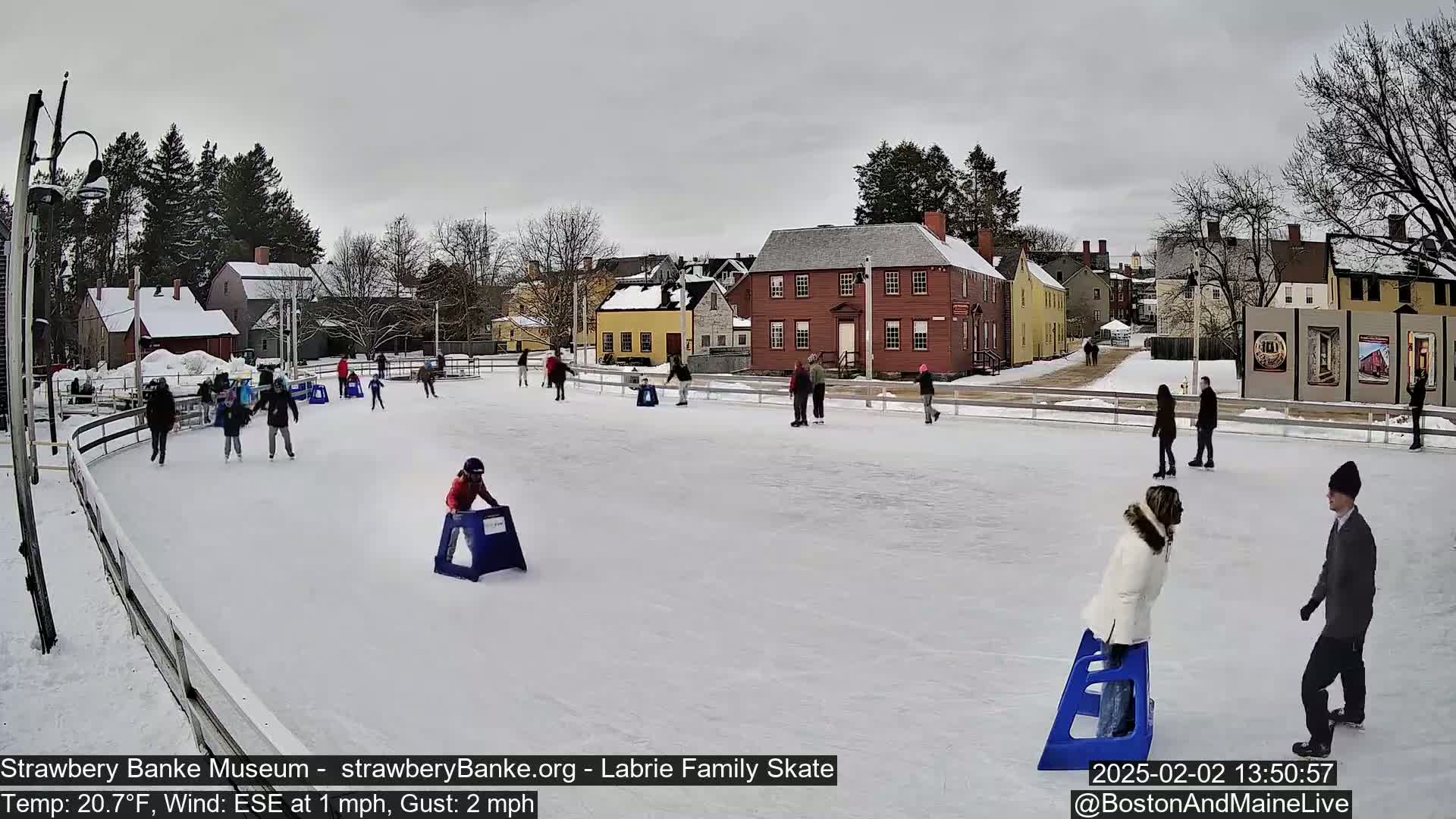 People ice skate on an outdoor rink in front of historical buildings under an overcast sky.