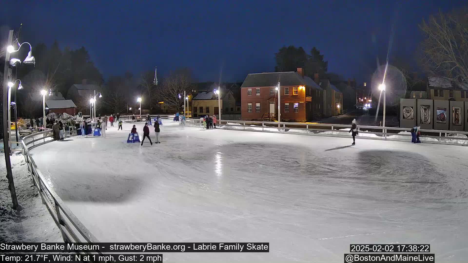 An outdoor ice-skating rink at night, illuminated by lights, is surrounded by buildings and trees under a dark sky.