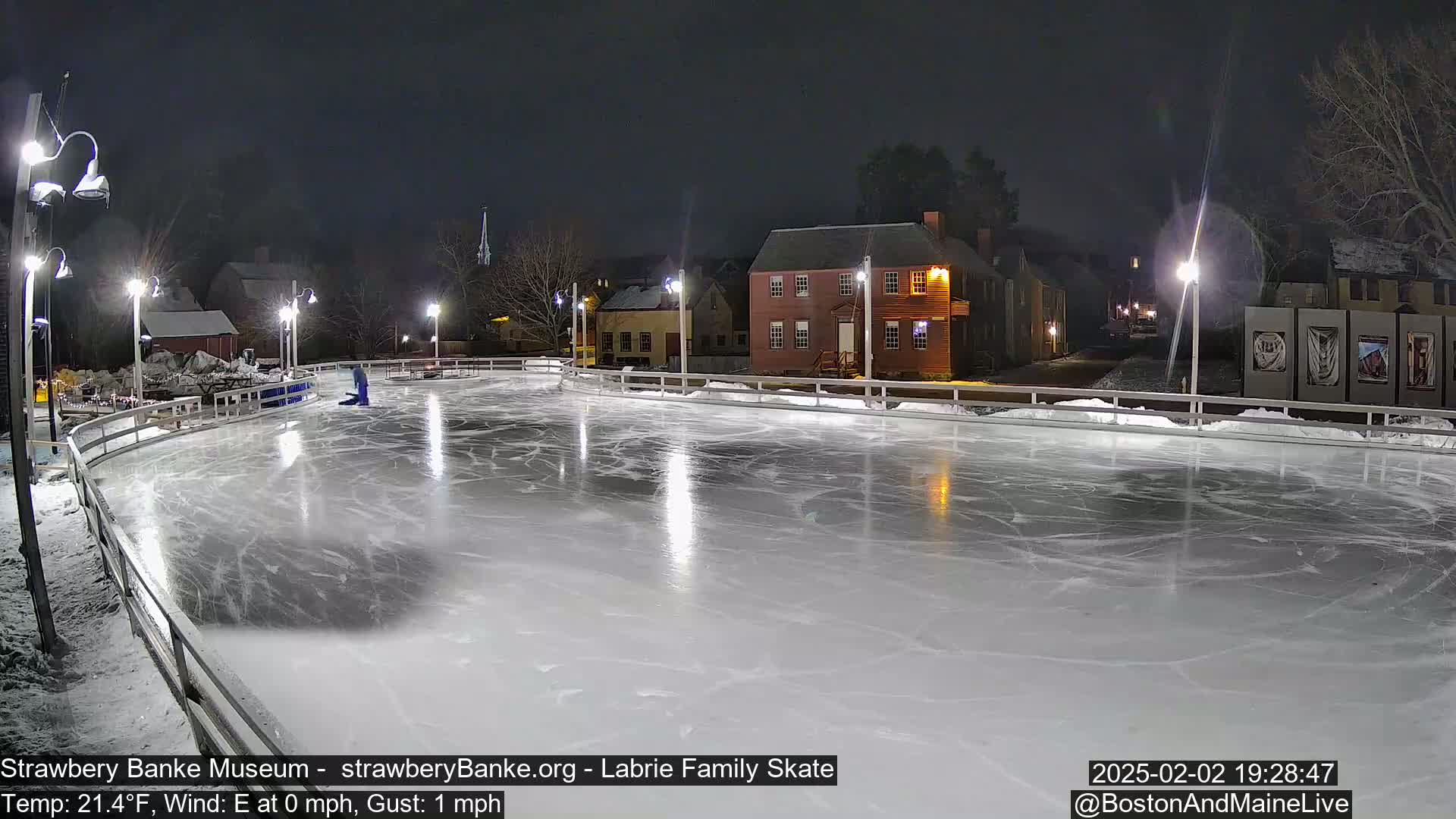 An outdoor ice skating rink at night, illuminated by lights, is surrounded by snow-covered buildings and trees under a dark sky.