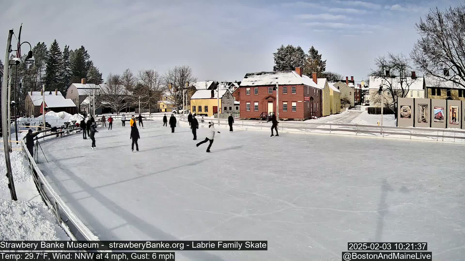 People ice skate on a rink outside in front of snow-covered buildings on a sunny day with a light wind.