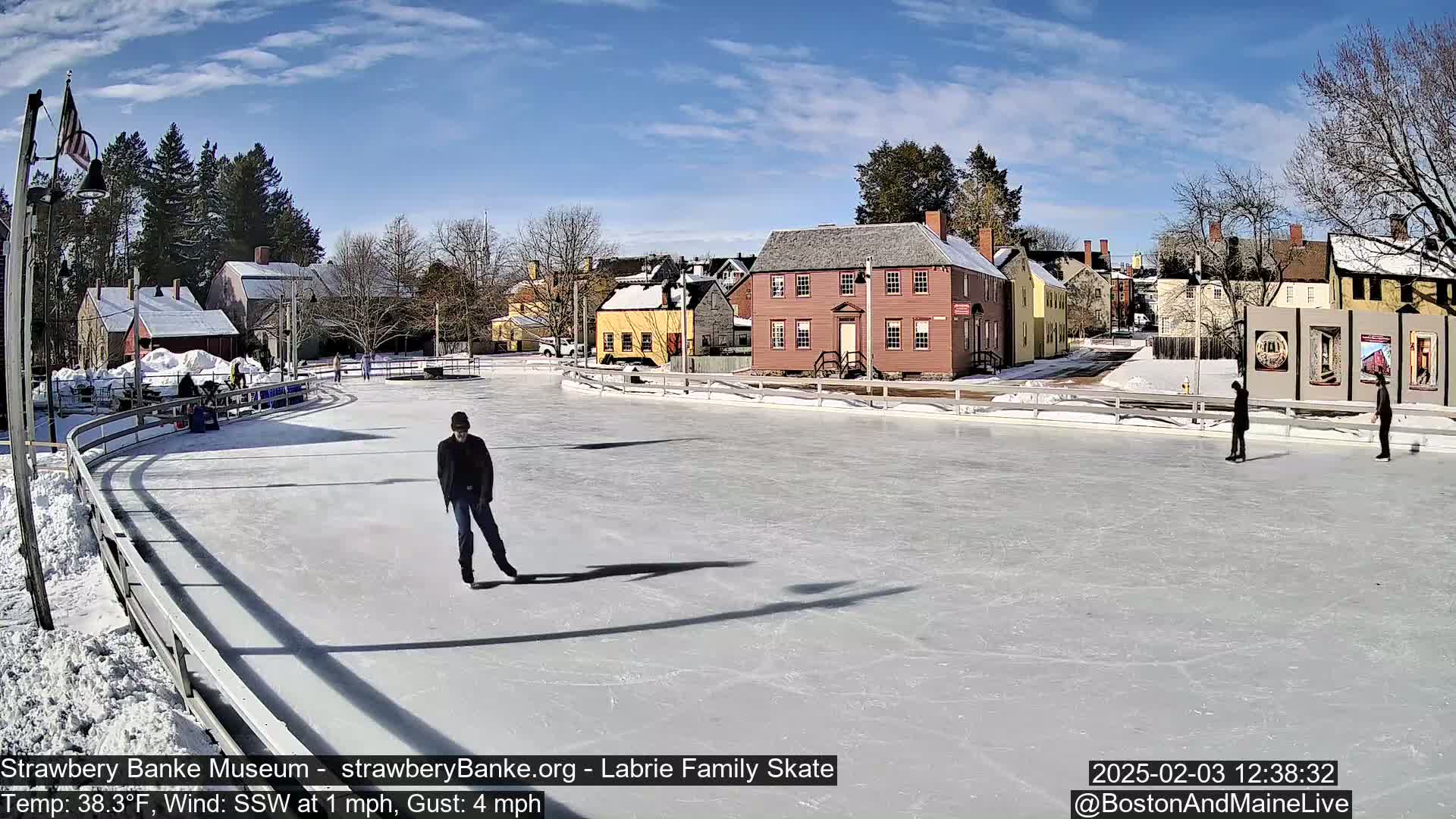 A sunny outdoor ice skating rink with several people skating, situated in front of a row of historic buildings.