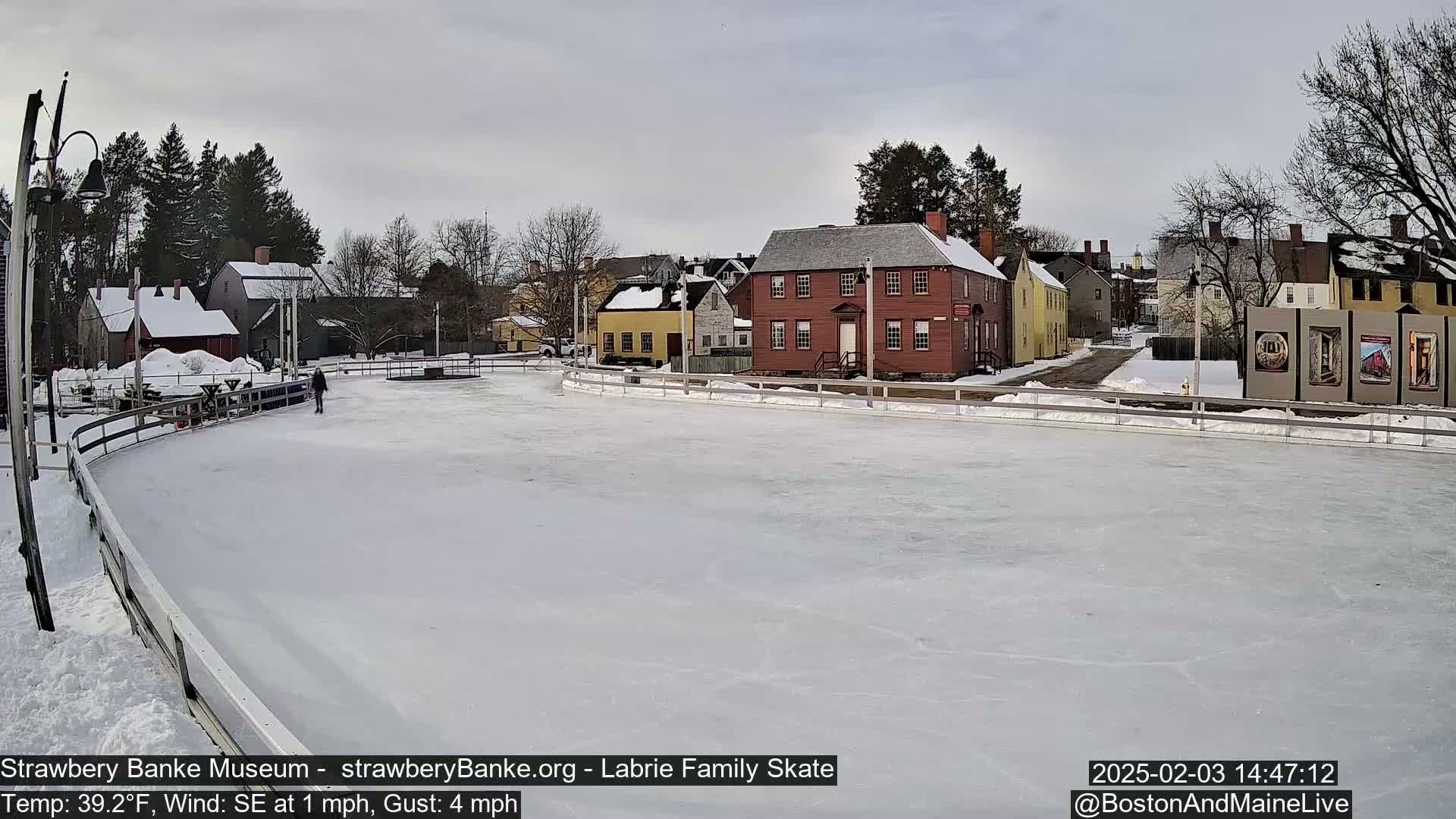 A person ice skates on an outdoor rink surrounded by snow-covered historic buildings under an overcast sky.