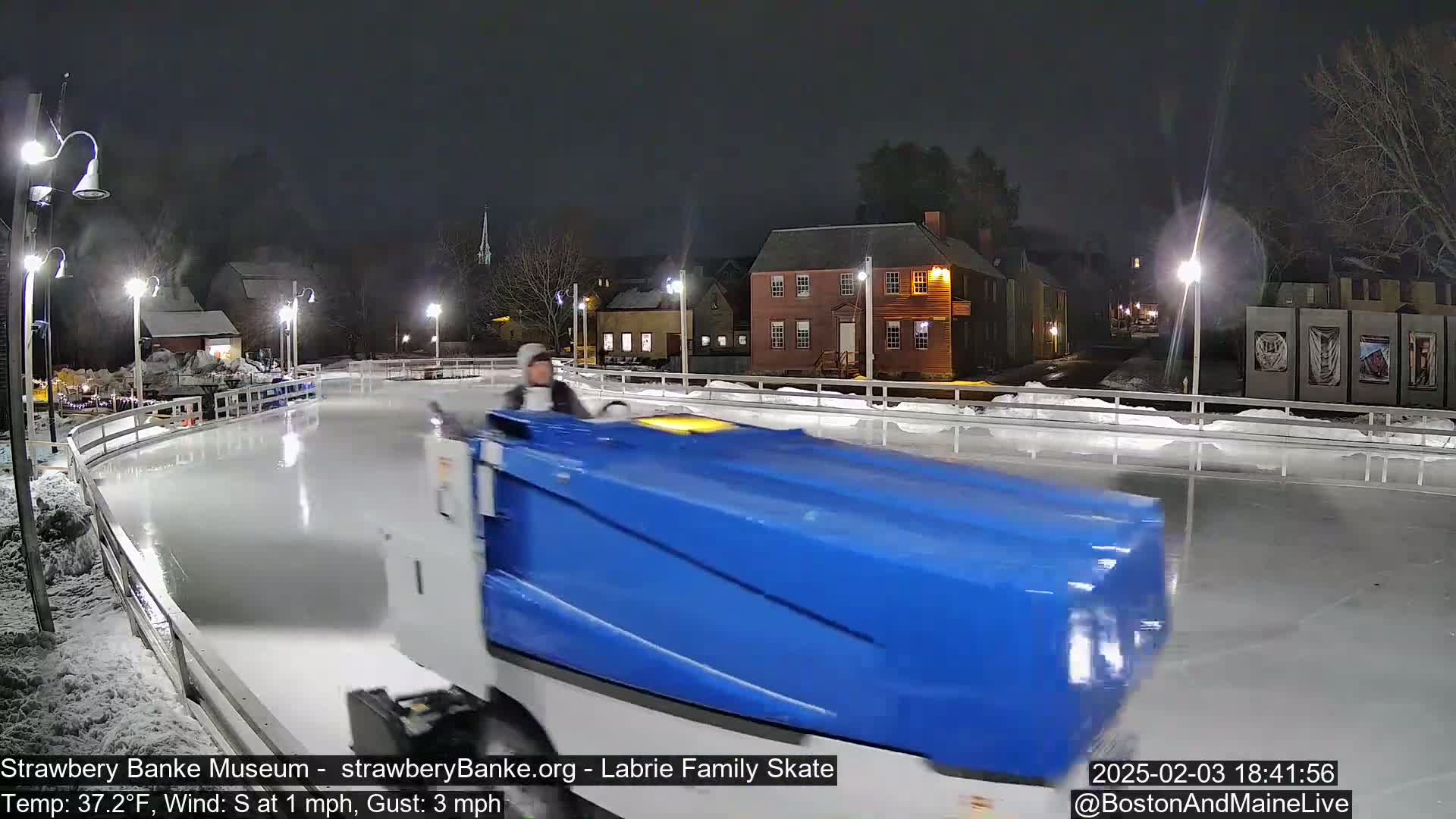 A blue Zamboni resurfacing an outdoor ice rink at night under dark skies.
