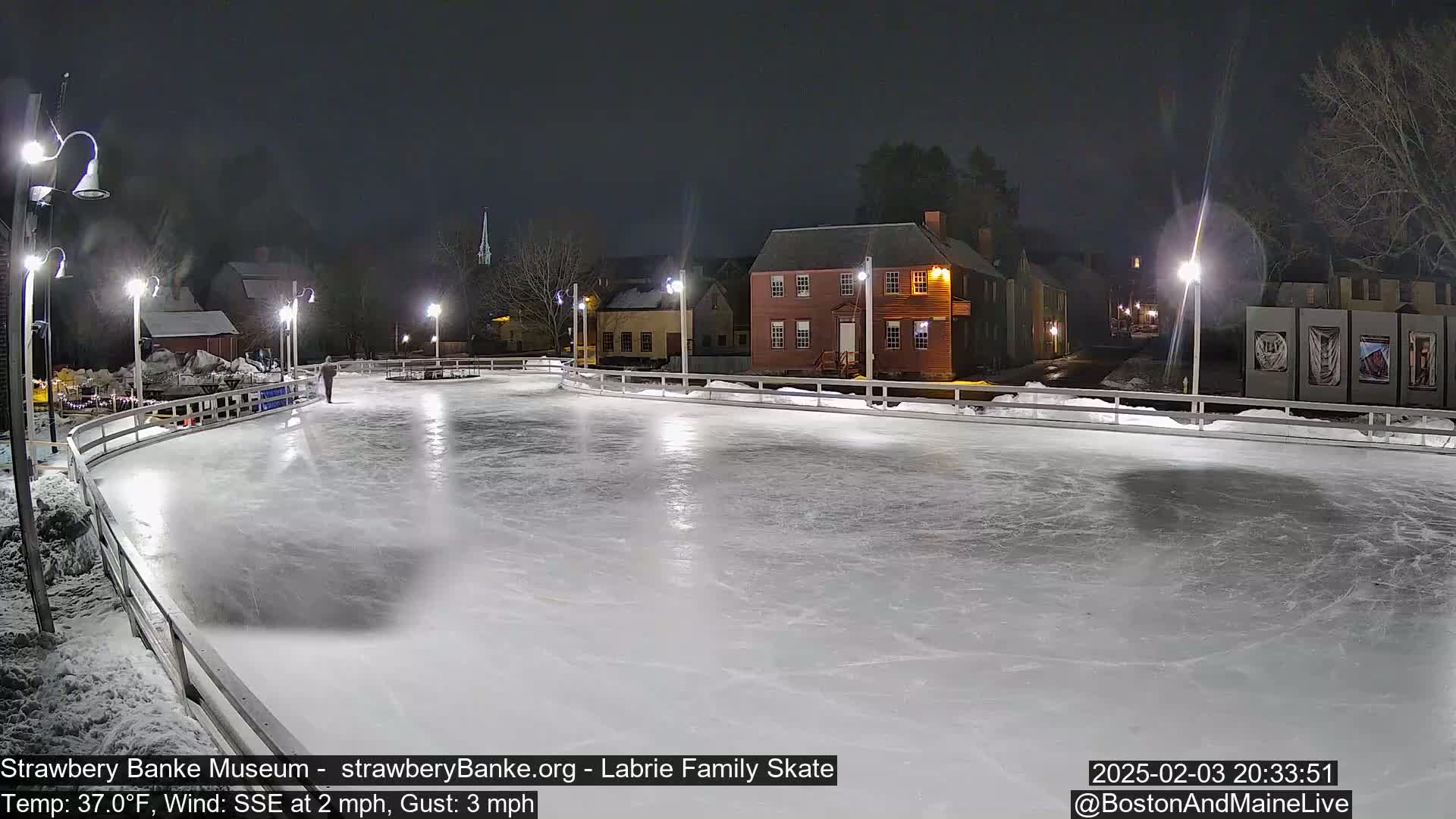 A nighttime view of an illuminated outdoor ice skating rink, with a single person skating and historic buildings in the background, under a dark sky with a light wind.