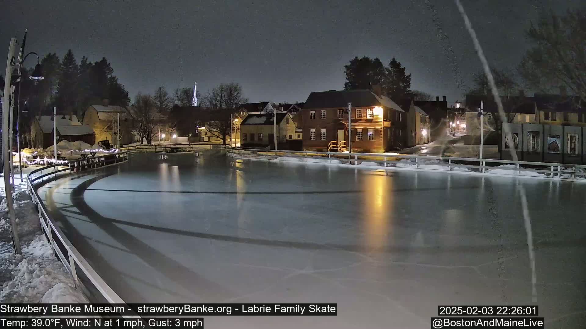 A nighttime view of an ice-skating rink illuminated by lights, surrounded by snow-covered buildings under a dark, possibly snowy sky.