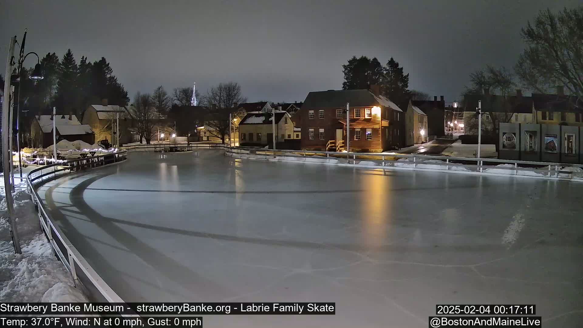 An outdoor ice skating rink at night, illuminated by nearby buildings, under a dark, overcast sky.