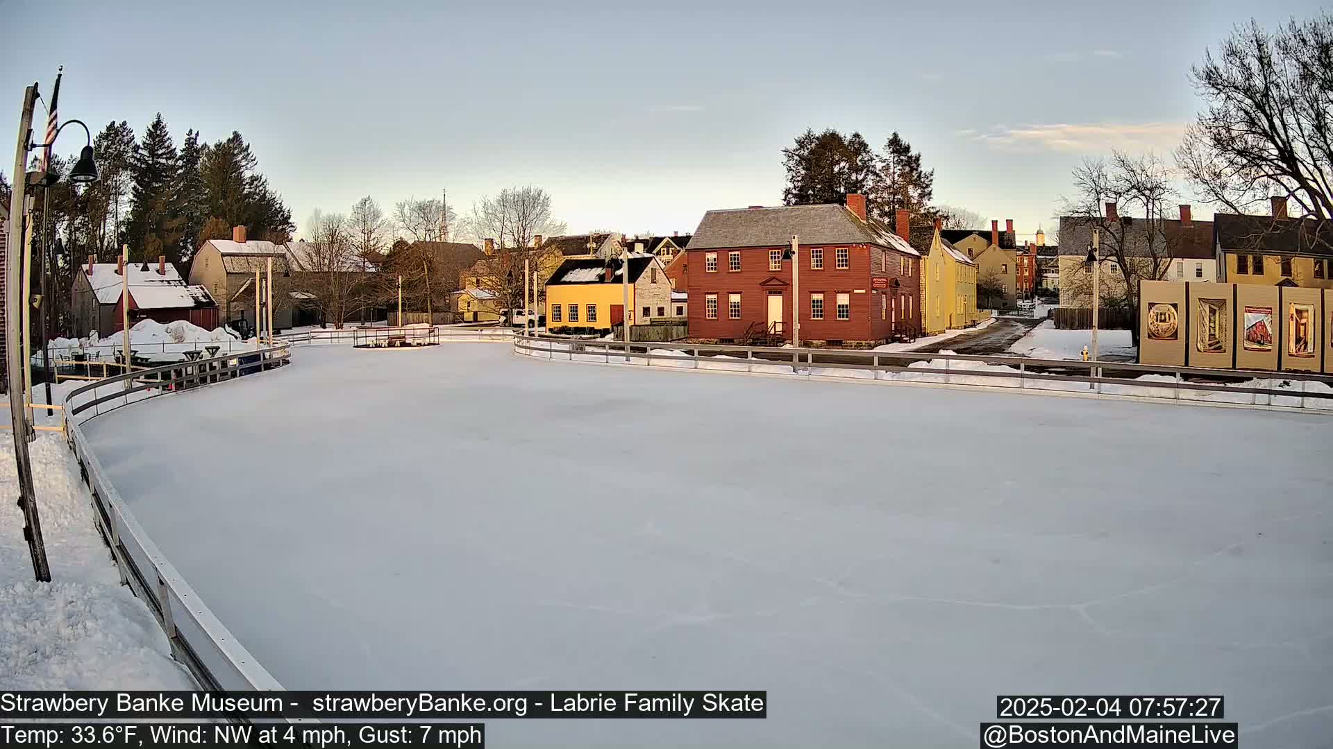 A snow-covered ice skating rink is situated in front of several historic-looking buildings under a clear, pale sky.