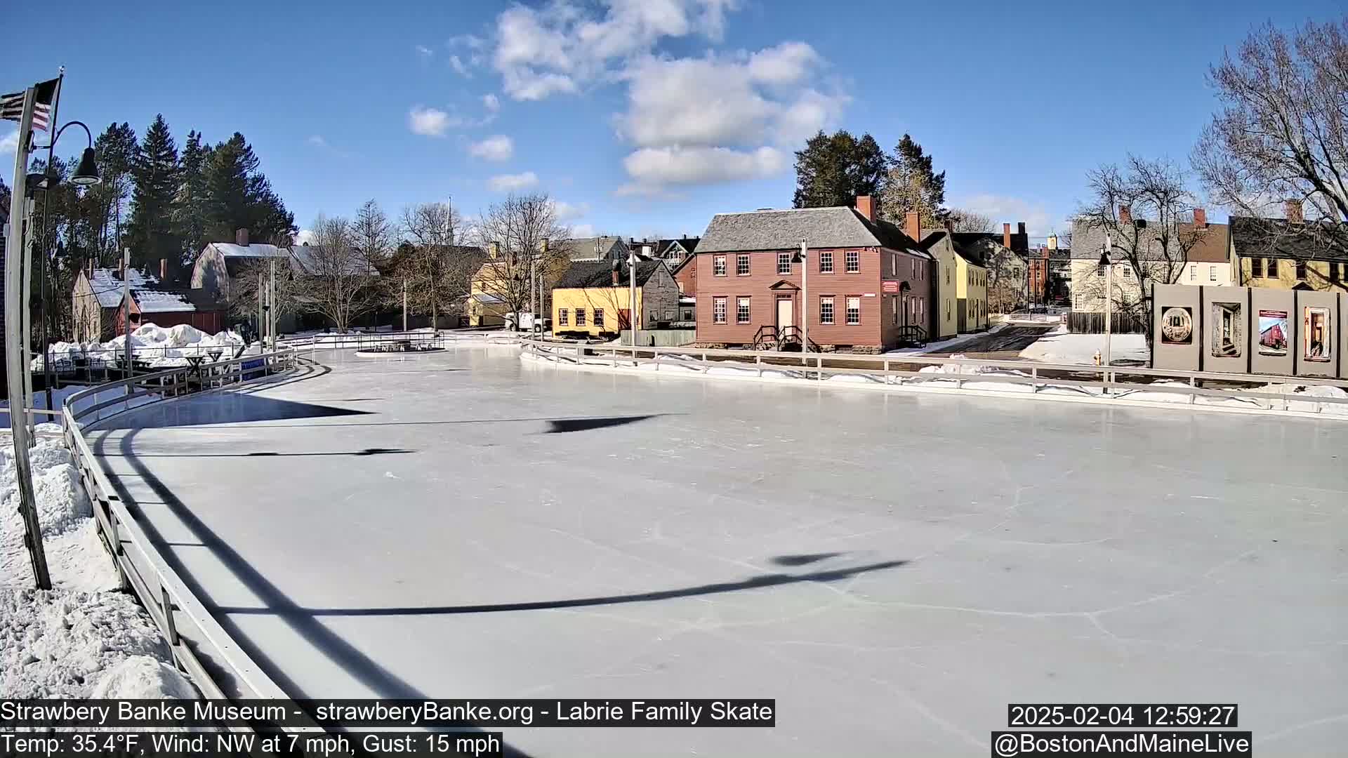 A frozen skating rink sits in front of several historic-looking buildings on a sunny day with a northwest wind.
