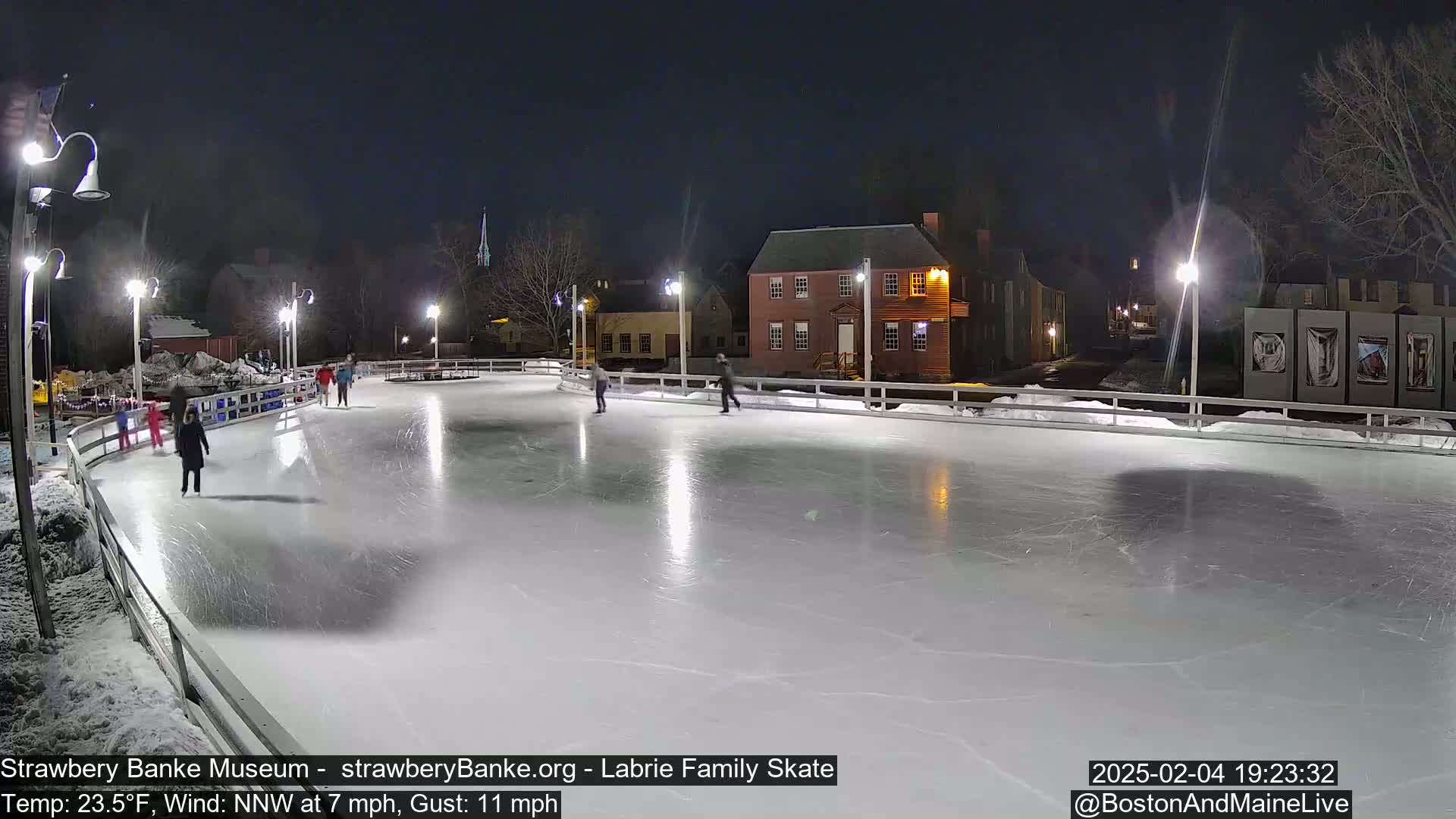 Several people ice skate on an outdoor rink at night under bright lights, with a backdrop of buildings and trees.