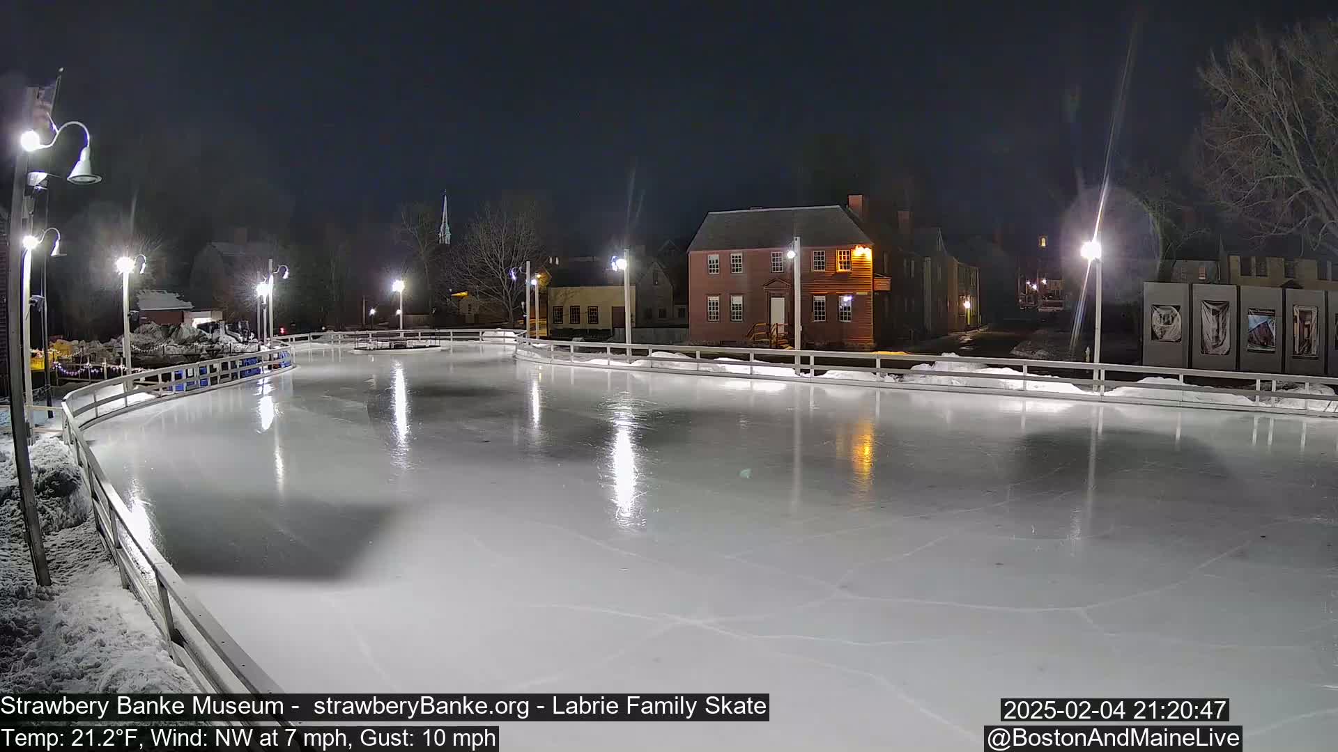 An outdoor ice skating rink, illuminated at night, is surrounded by buildings and trees under a dark sky with a northwest wind.